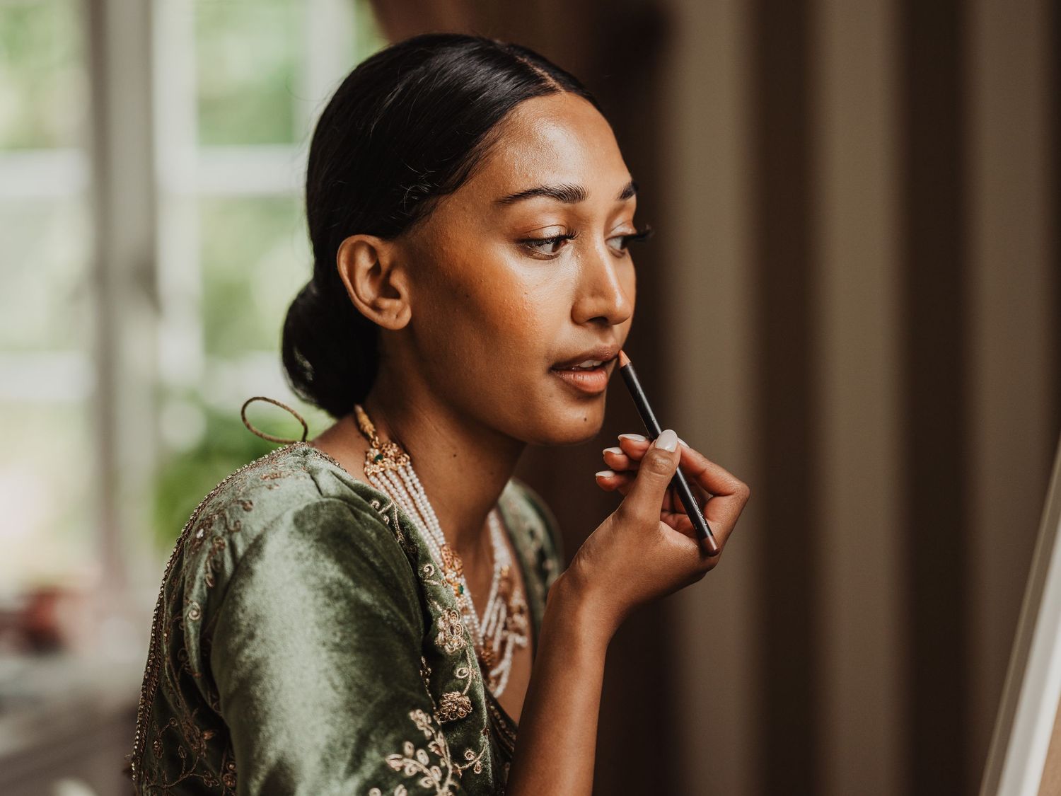 Beautiful South Asian bride applying lipstick while getting ready in a green embroidered bridal outfit.