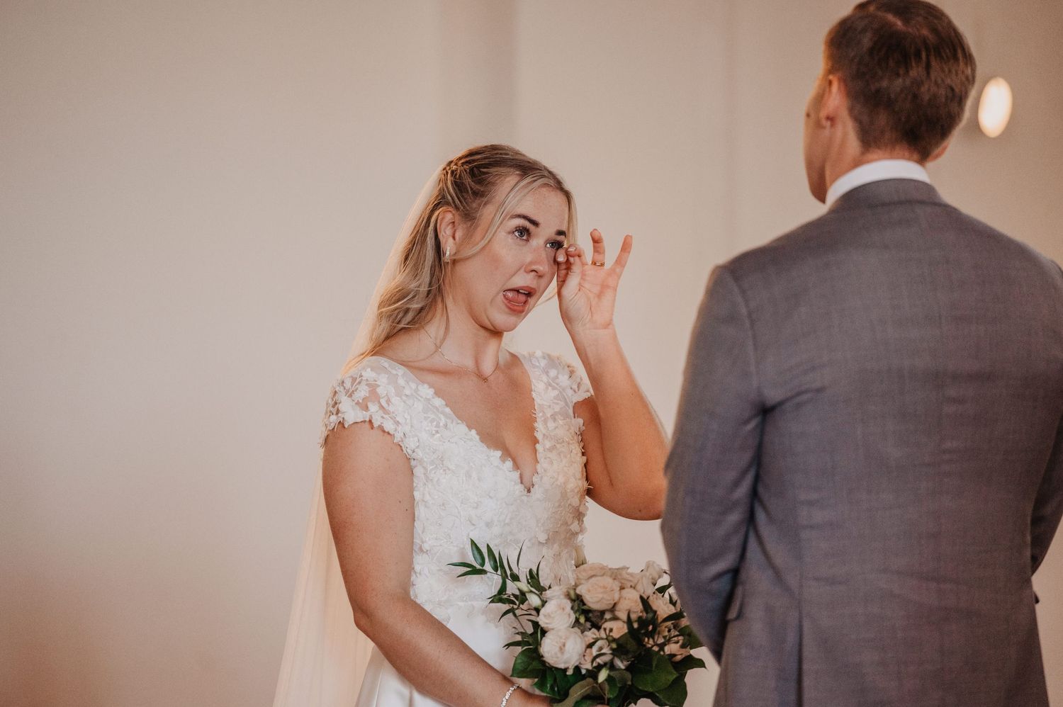 Emotional bride in lace wedding dress wipes tears while holding bouquet during heartfelt moment.