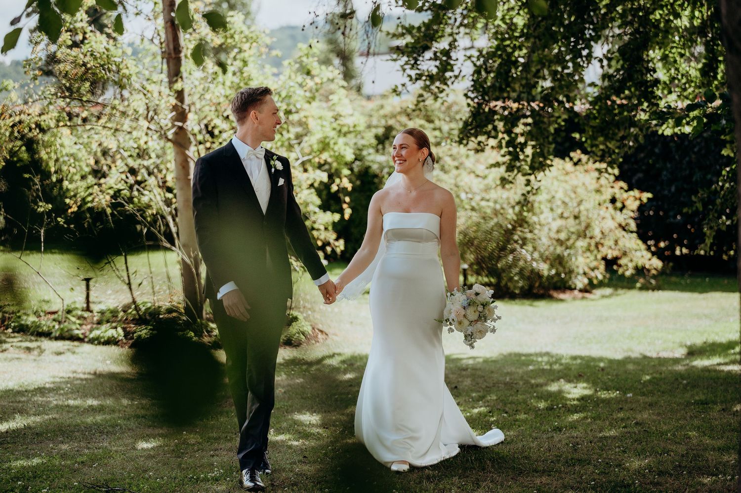 A bride and groom walk hand in hand through a lush green garden on their wedding day.