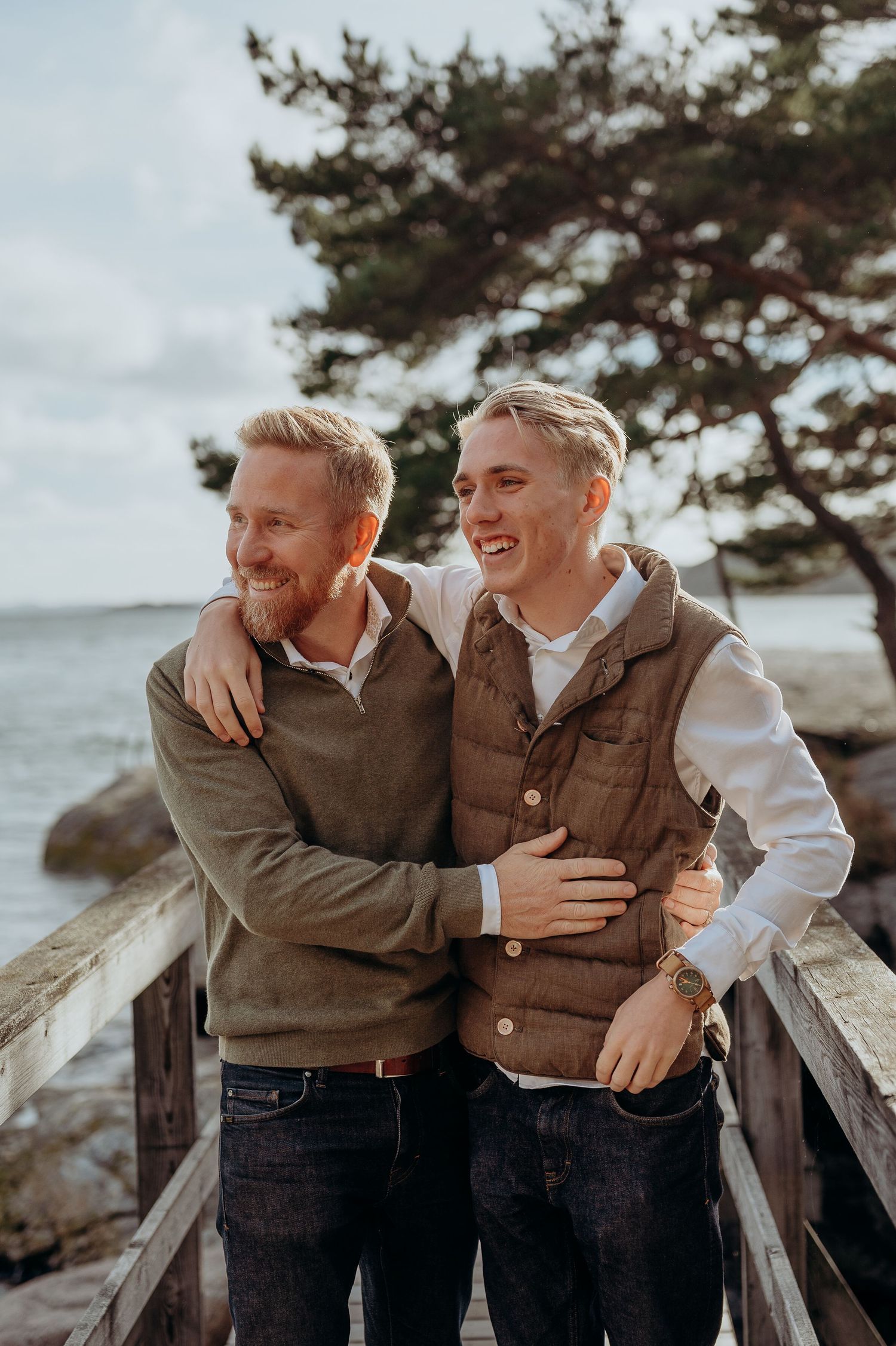Two smiling men embrace on a wooden bridge by the sea, wearing casual autumn outfits.