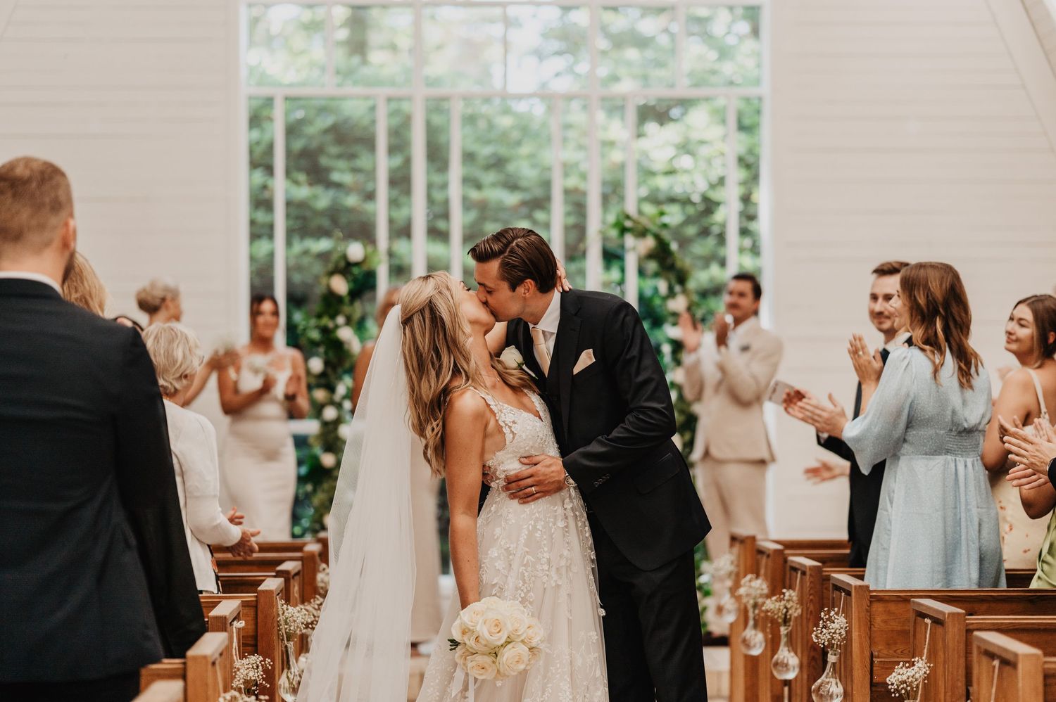 Bride and groom share first kiss at church altar while guests applaud during elegant indoor wedding ceremony.