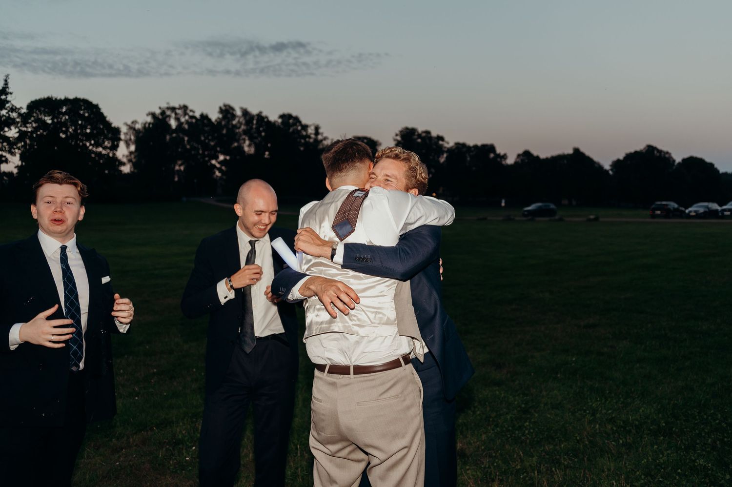 A groom shares a heartfelt embrace with a guest at an outdoor evening wedding celebration.