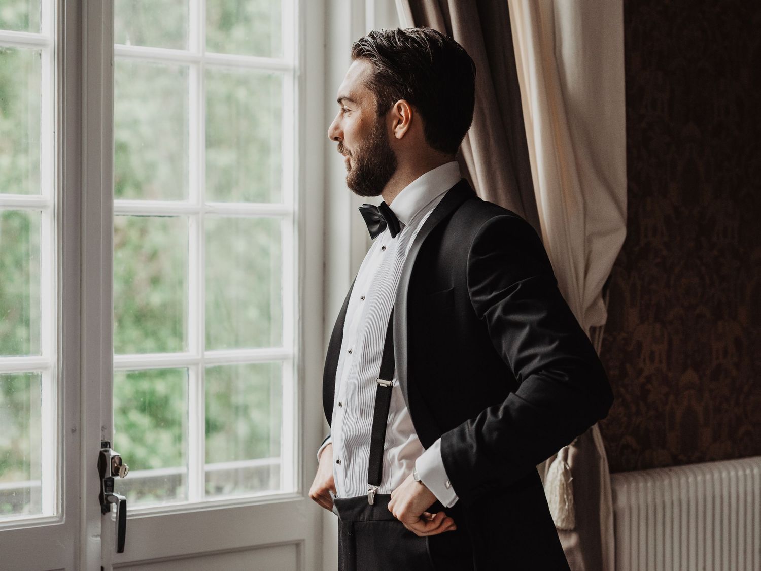 An elegant groom in a black tuxedo with bow tie stands gazing out a large window before his wedding.
