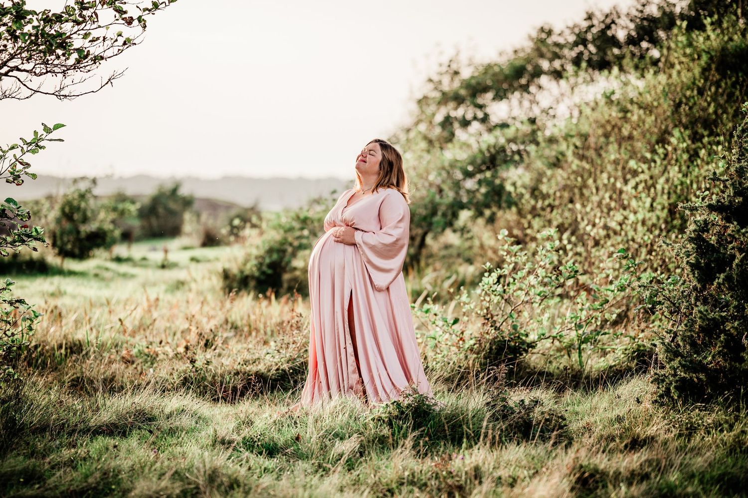 Pregnant woman in pink flowing dress standing gracefully in a lush green hillside landscape.