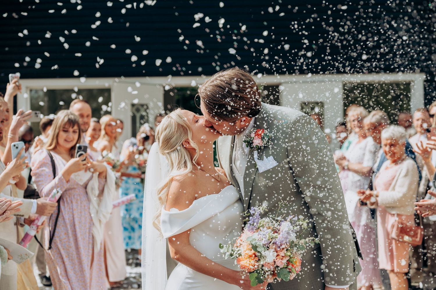 Newlyweds kissing in a shower of white confetti surrounded by joyful guests outdoors.