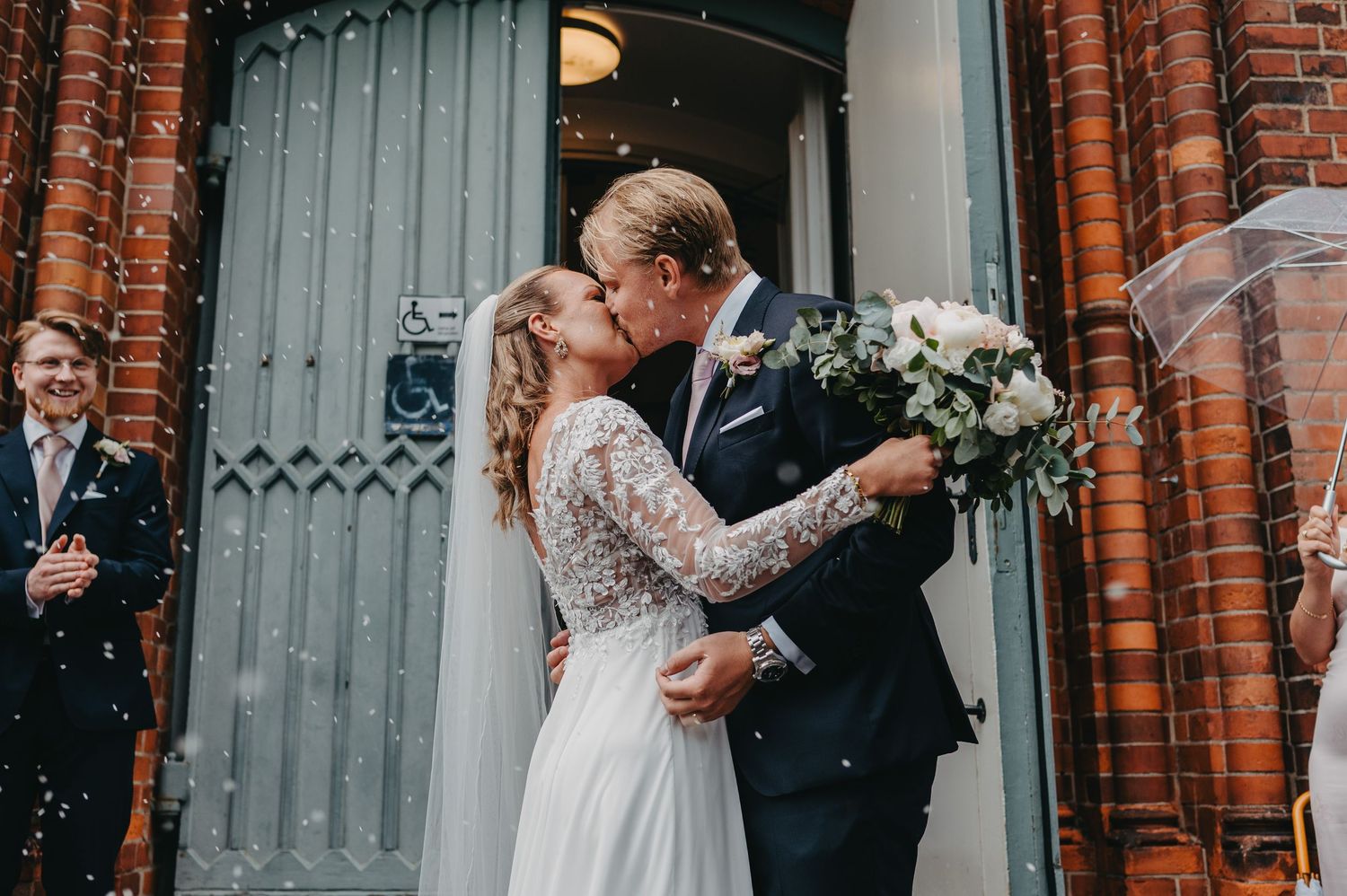 Newlywed couple sharing a romantic kiss outside church doors with confetti falling, bride holding white bouquet.