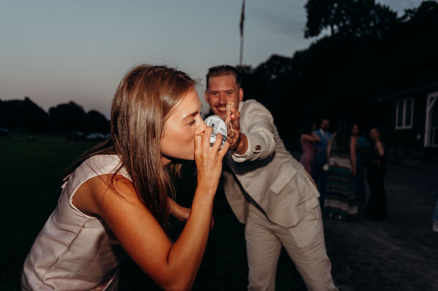 A woman kisses a wedding favor while a man in a white suit reaches out at an outdoor evening wedding reception.