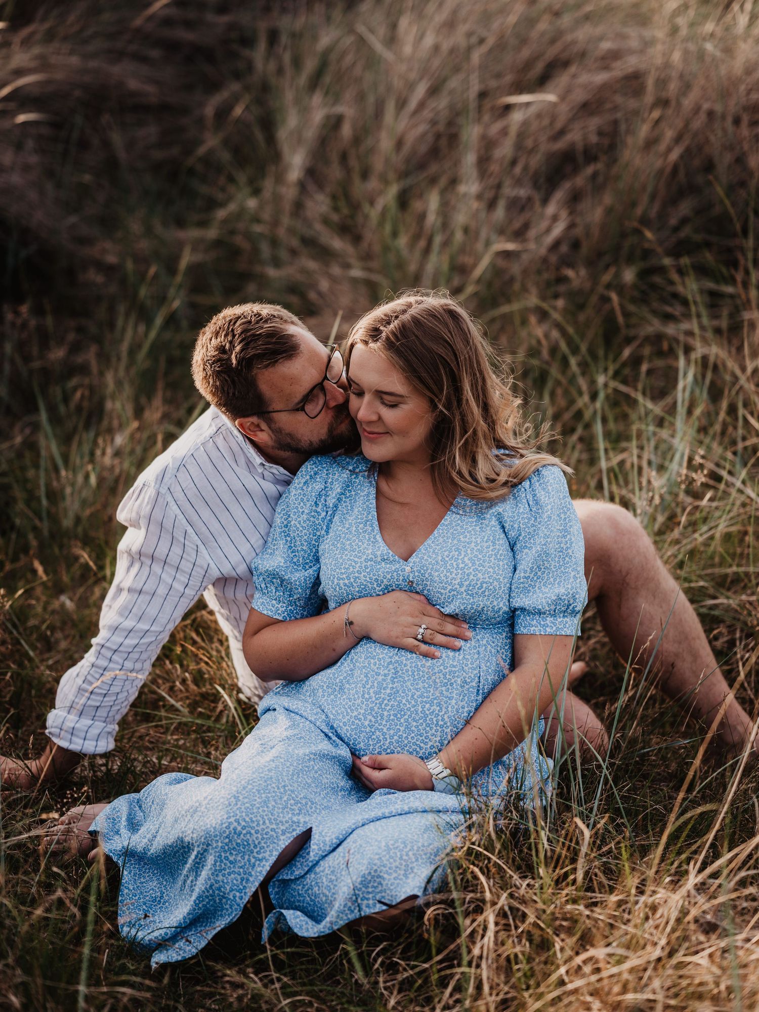 Couple sitting in tall grass as man kisses pregnant womans cheek in a field.