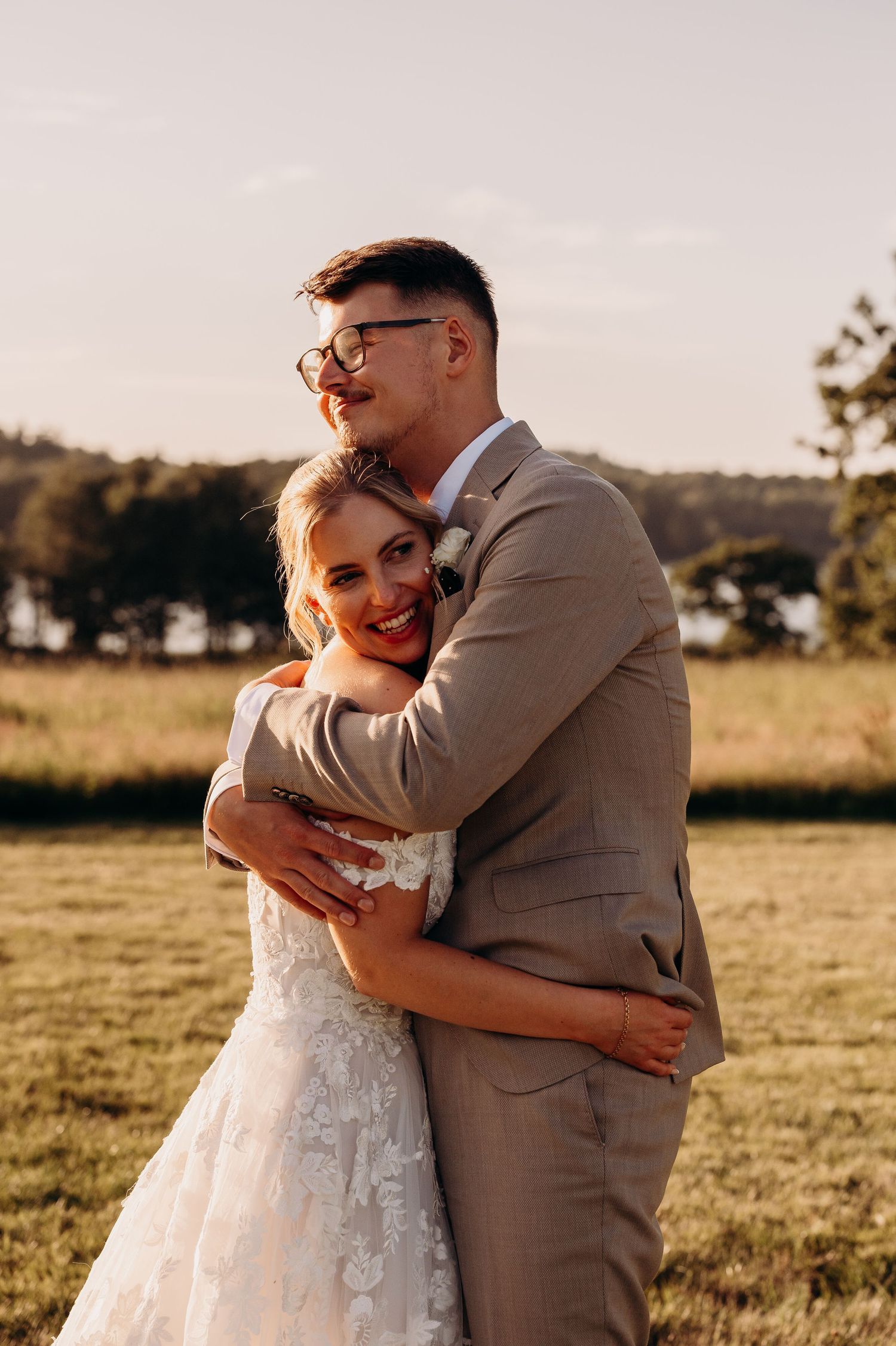 A smiling bride in a lace gown embraces her groom in a tan suit outdoors during golden hour.