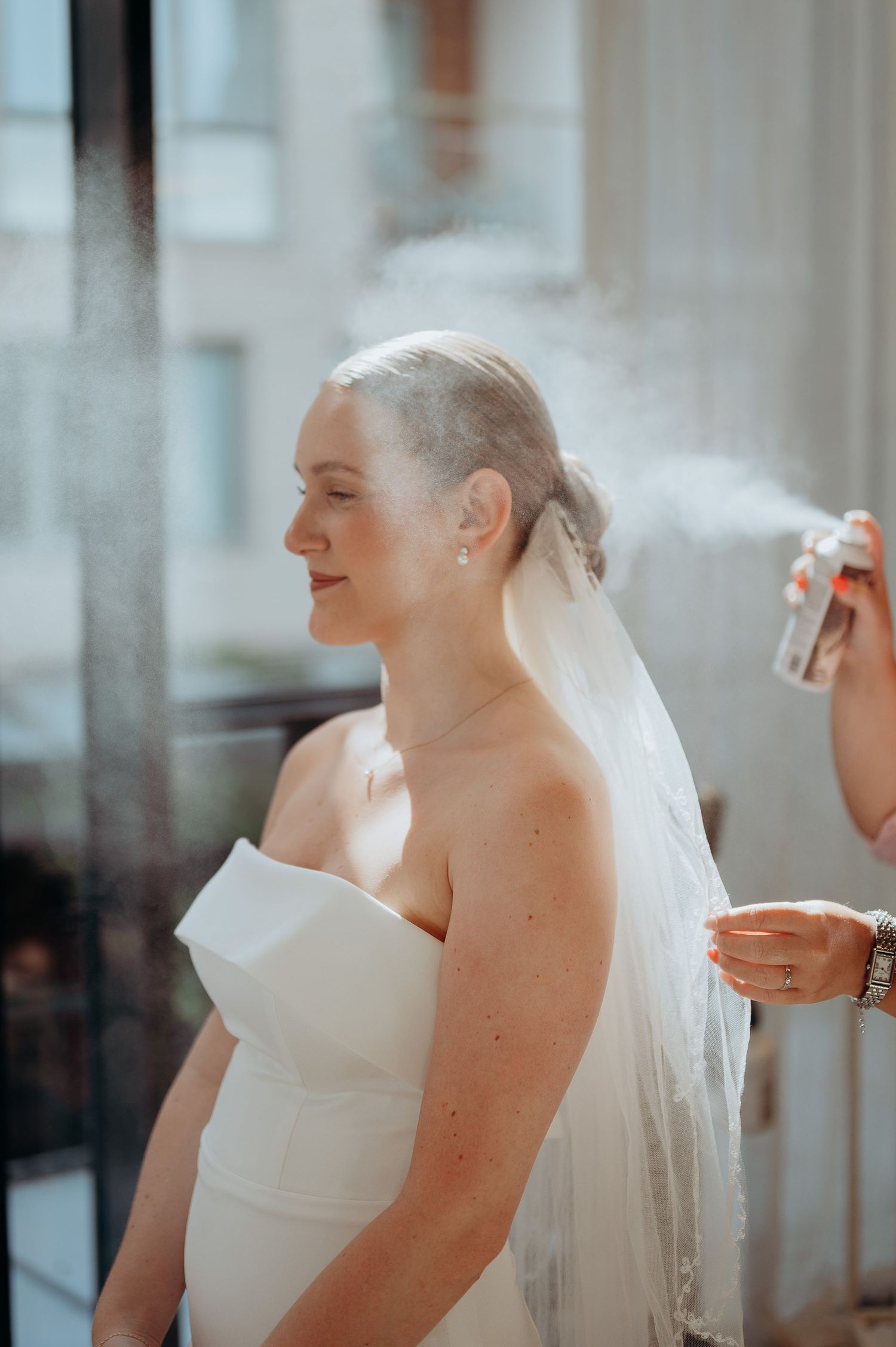 Bride in strapless white gown with veil gets hairspray applied during wedding preparations indoors.