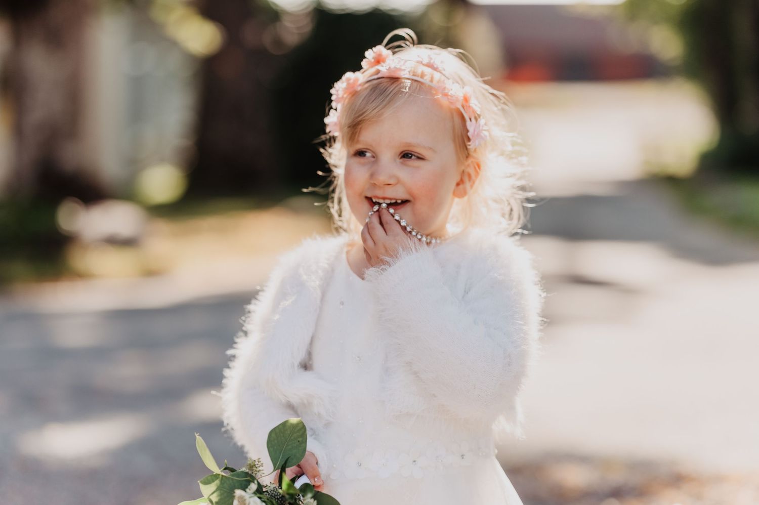 Adorable little girl in white dress and floral headband holds bouquet outdoors.