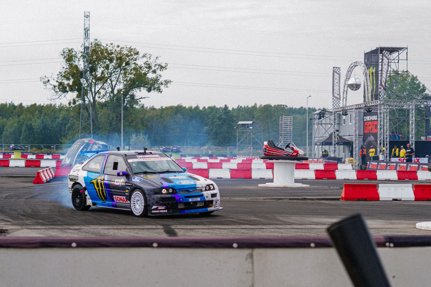 Drift racing cars perform stunts on a track with red and white barriers.
