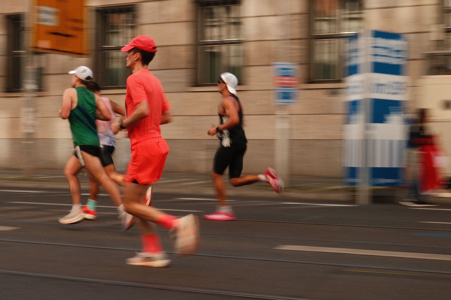 Athletes in red and green running gear sprint down an urban street during a competitive race event.