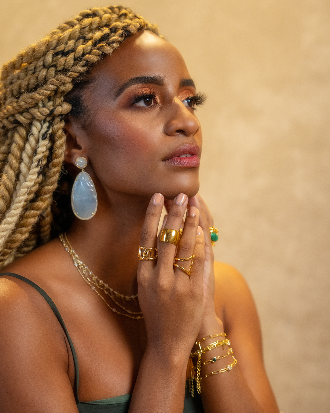 Blonde braids and statement jewelry featured in warm-toned portrait against beige background.