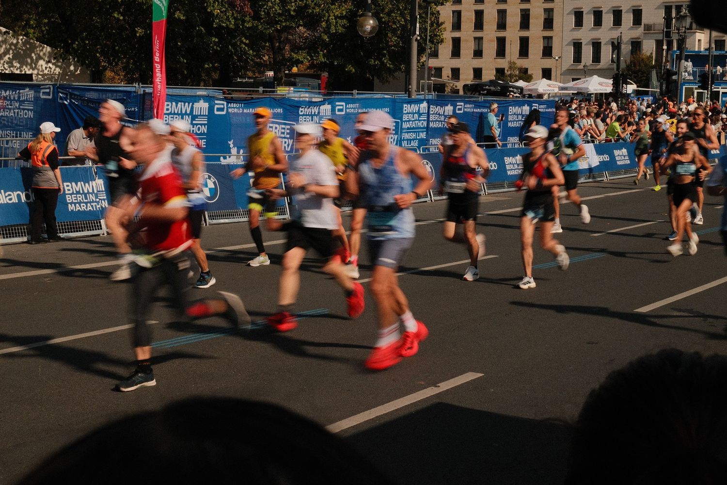 Marathon runners compete in a city road race on a sunny day with blue barriers lining the course.