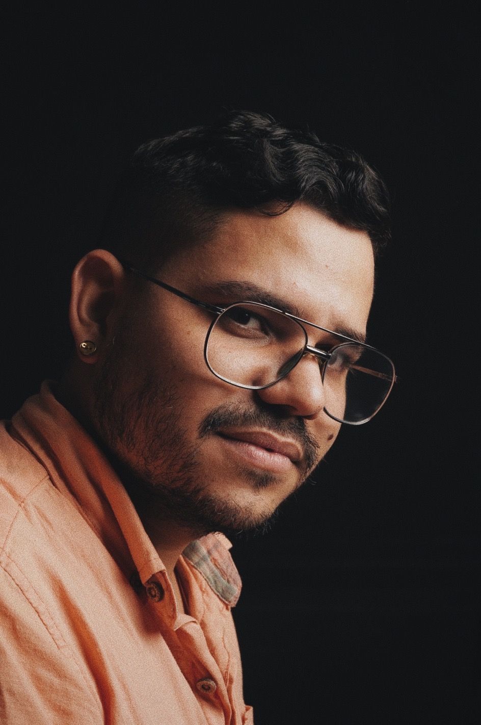 Portrait of someone in an orange shirt against a dark background wearing glasses.