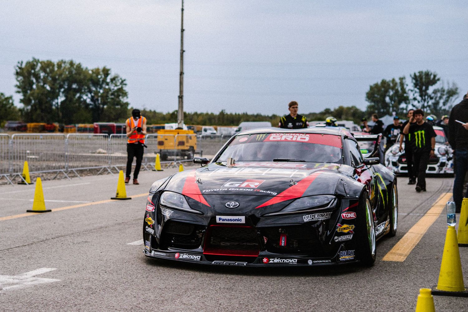 Race cars line up on a track with yellow safety cones and barriers.