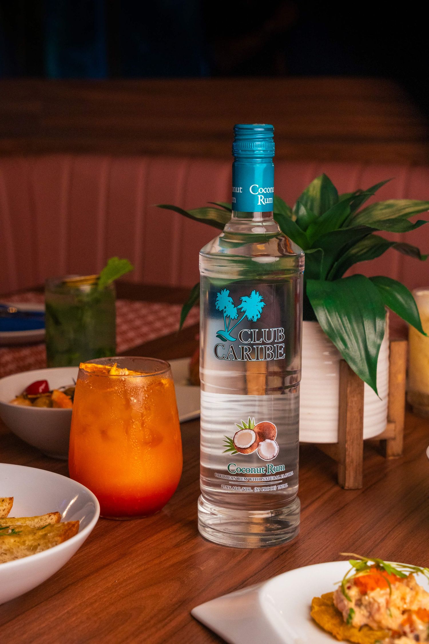 Restaurant table setting with tropical drinks and a bottle of Club Caribe clear spirits against wooden backdrop.