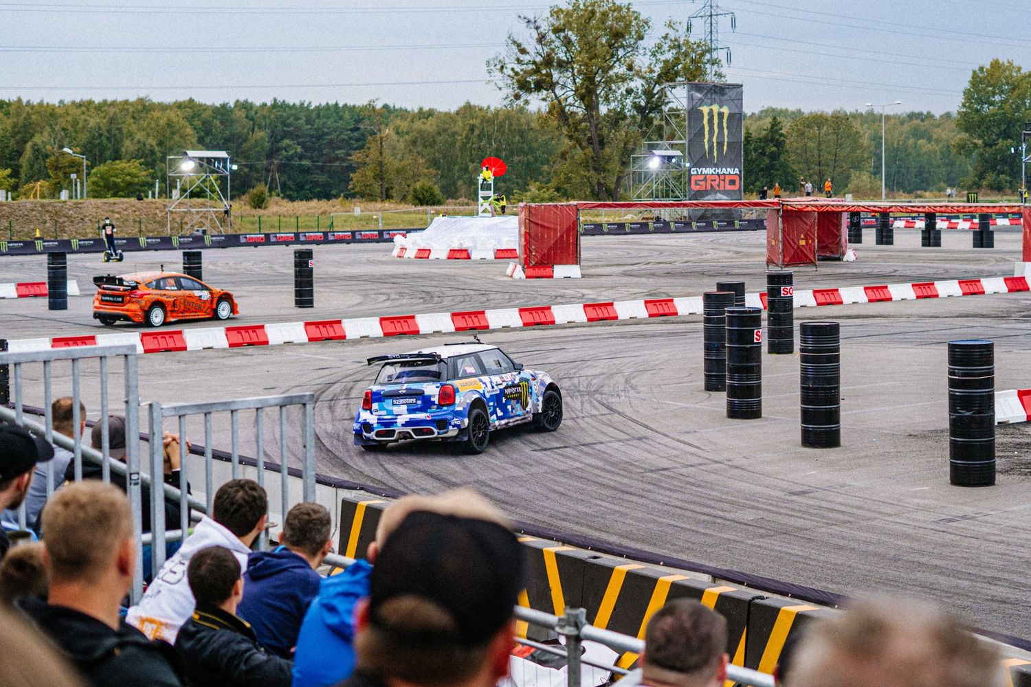 Spectators watch drift racing cars perform on a track with red and white barriers.