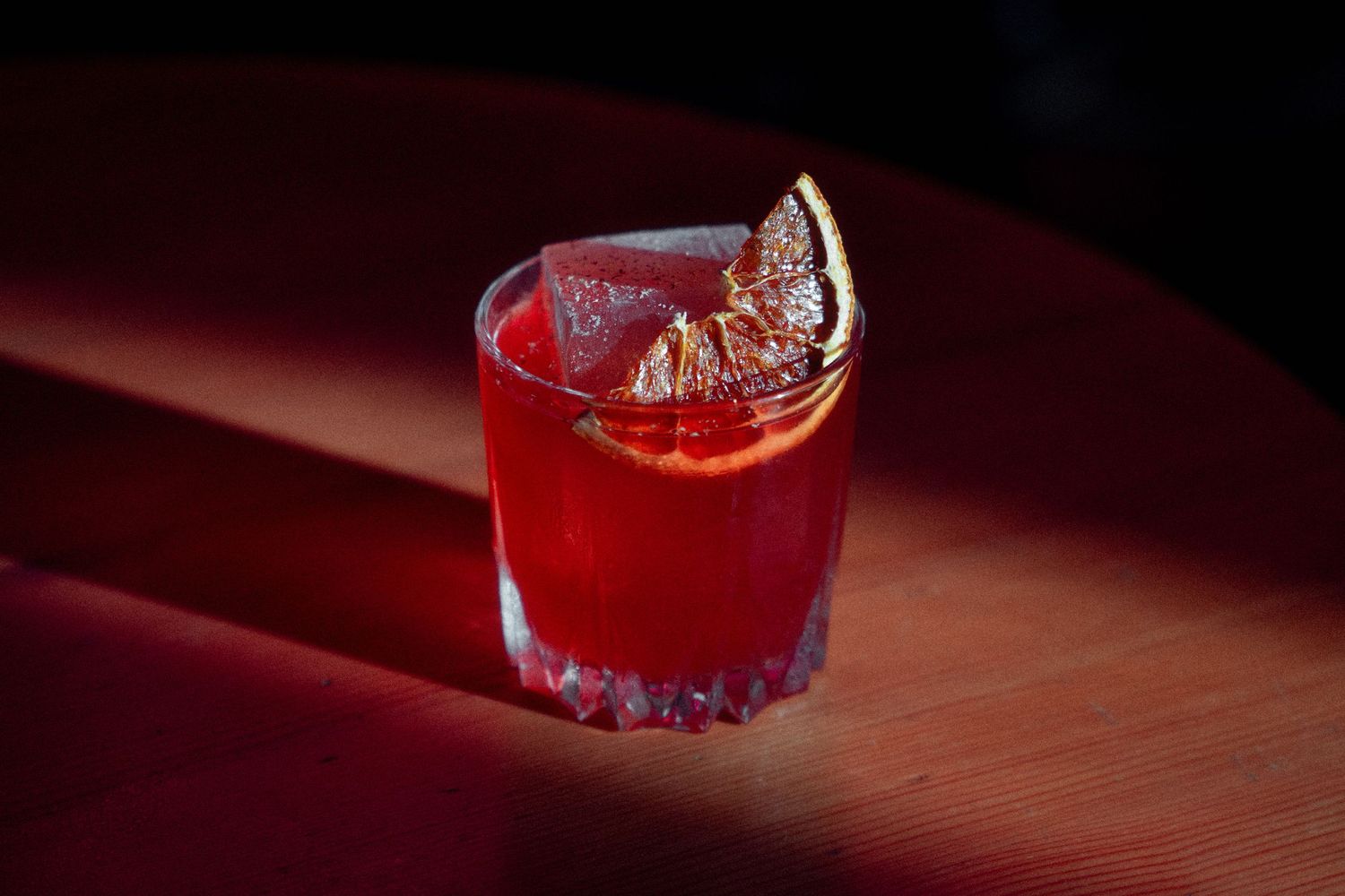 Red cocktail in crystal glass on dark wooden surface with moody lighting.