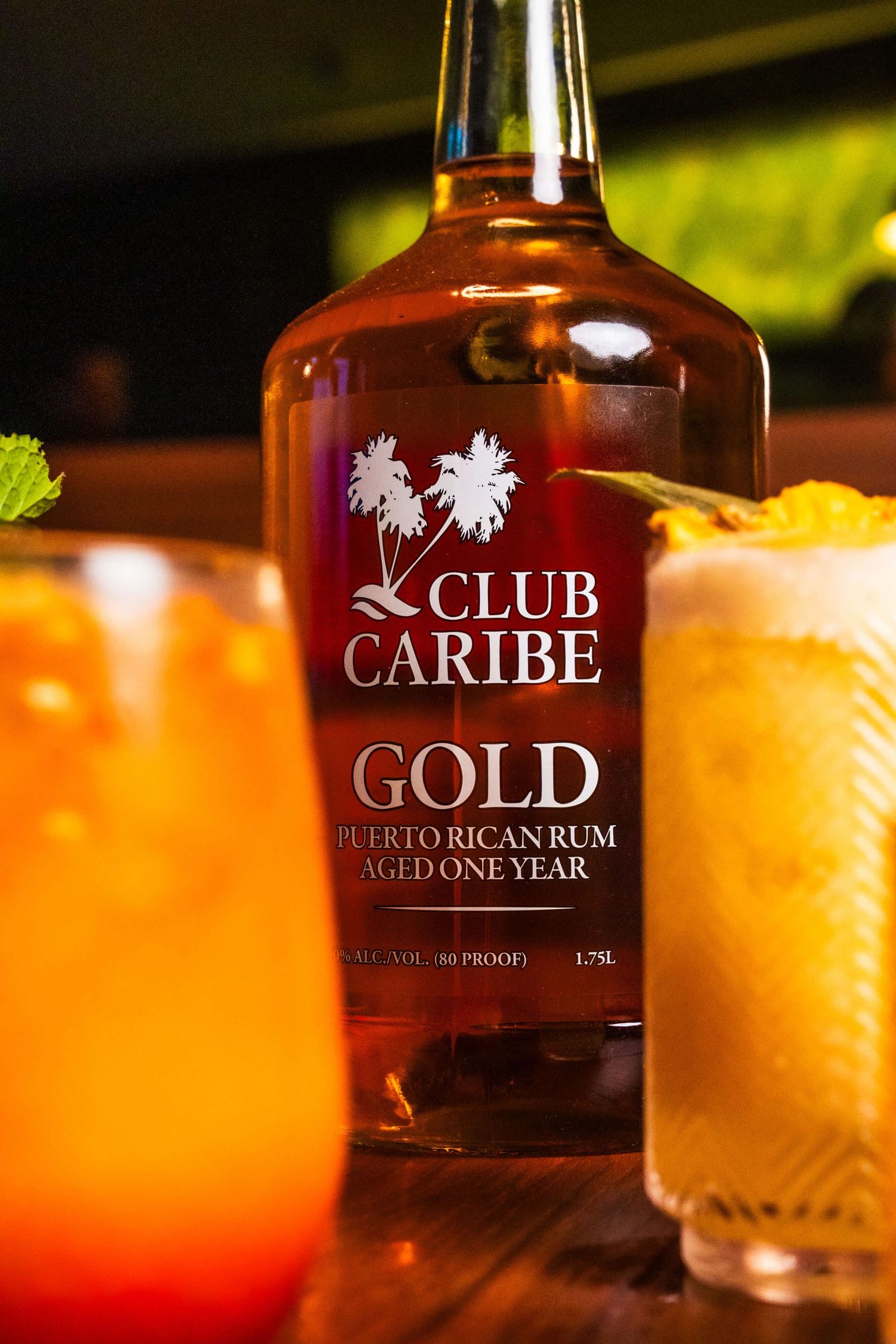 Row of tropical cocktails with Club Caribe Gold rum bottles in warm ambient lighting.