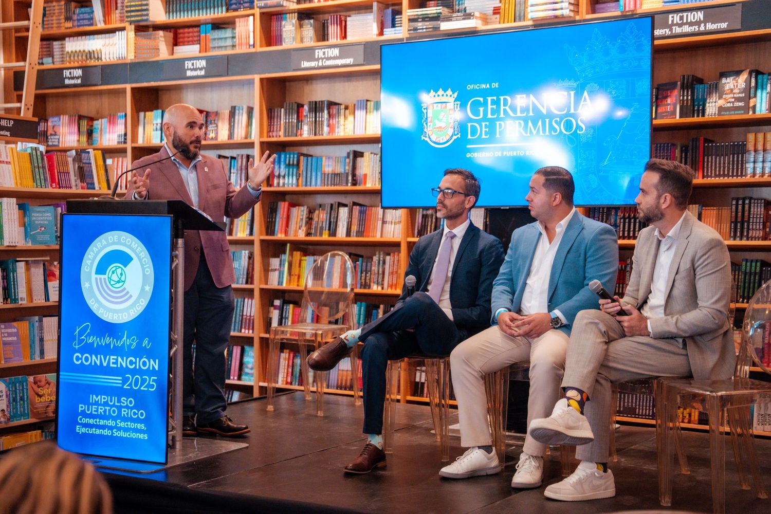 Panel discussion taking place in a library with bookshelves and blue banners in the background.