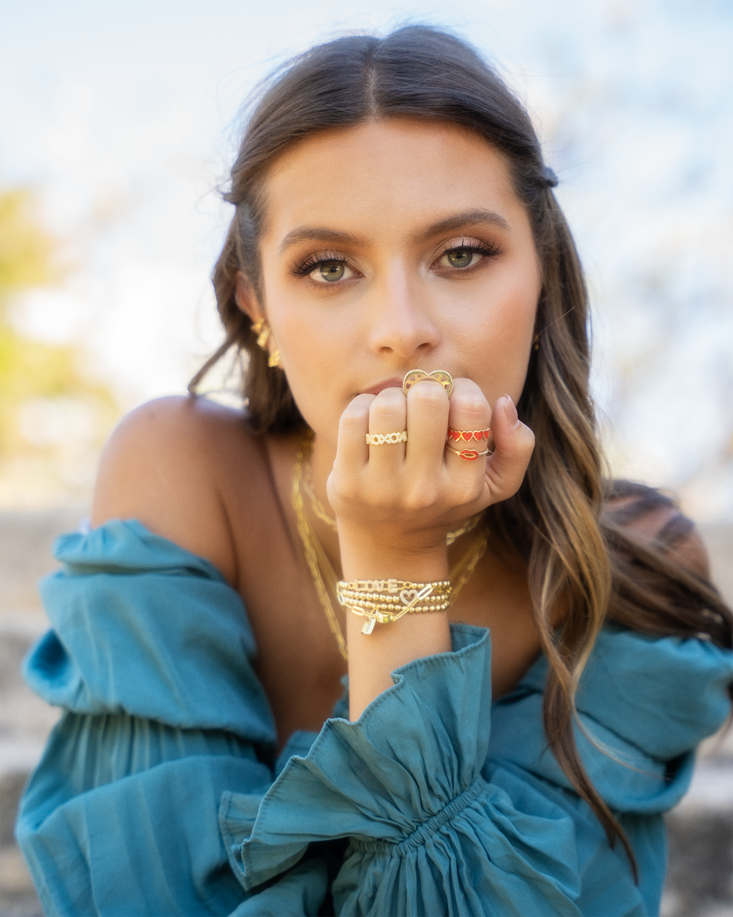 Fashion portrait featuring teal ruffled dress and layered gold jewelry against blurred background.