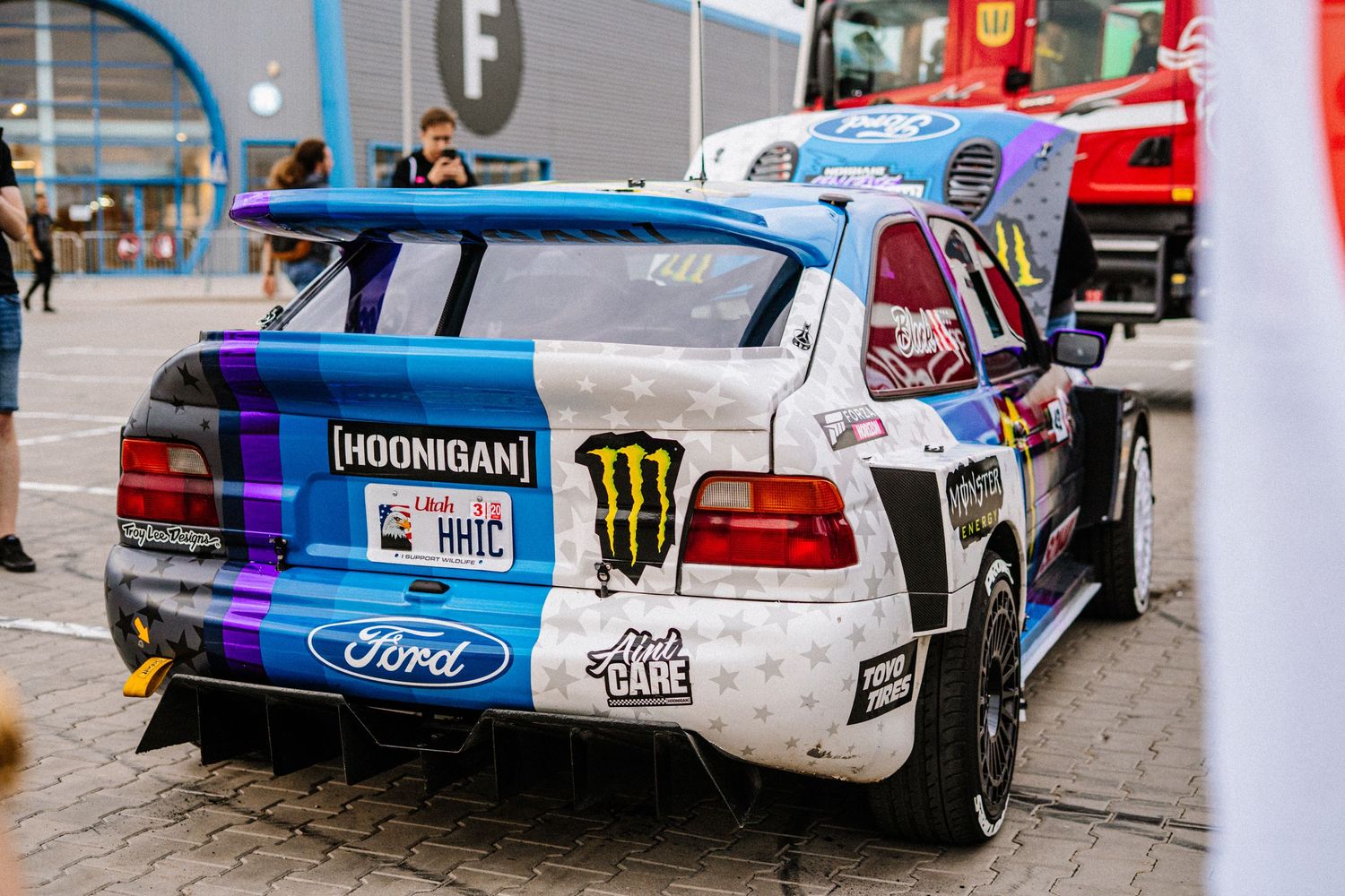 Modified racing cars with sponsor decals parked in a row at a motorsport event.