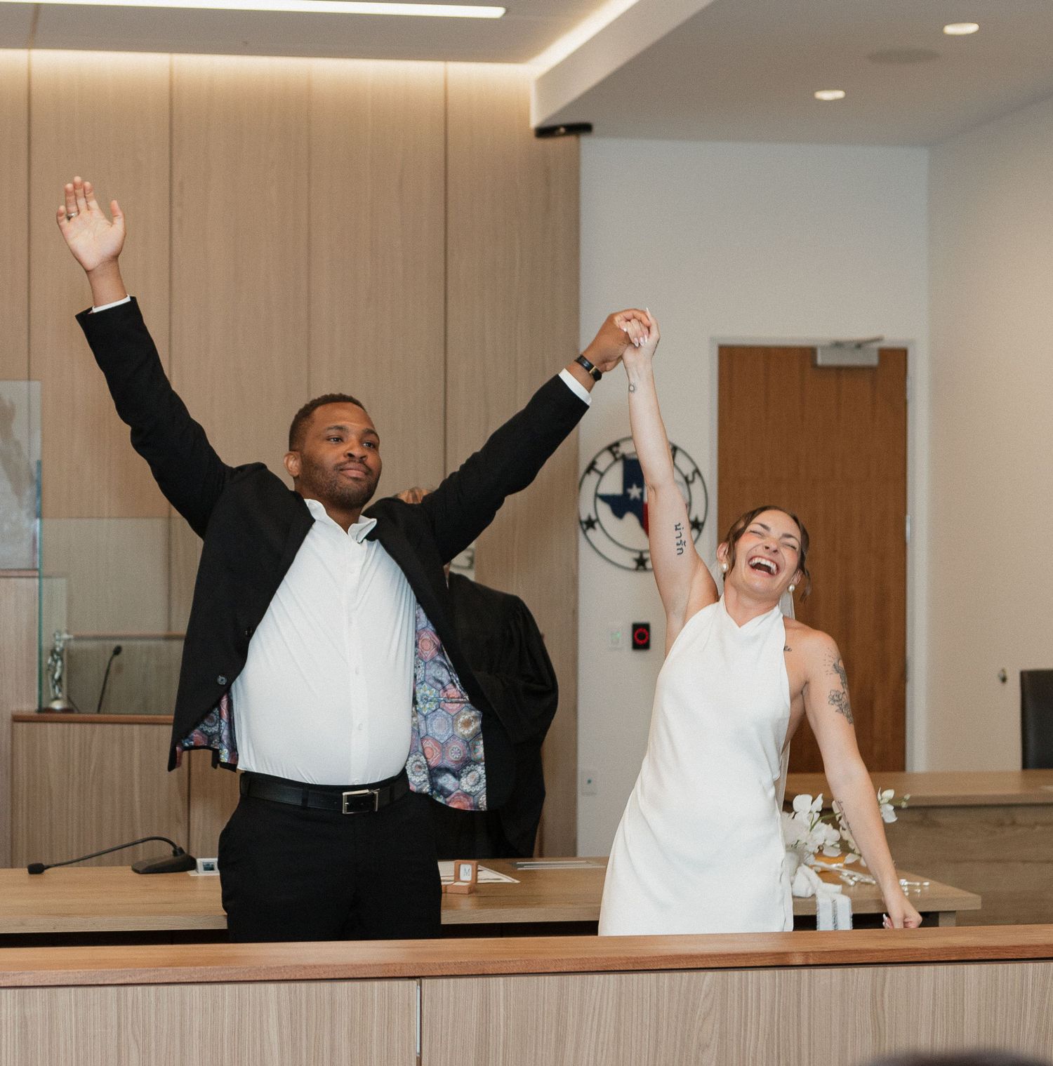 A couple in wedding attire jumps and celebrates joyfully in a modern indoor setting.