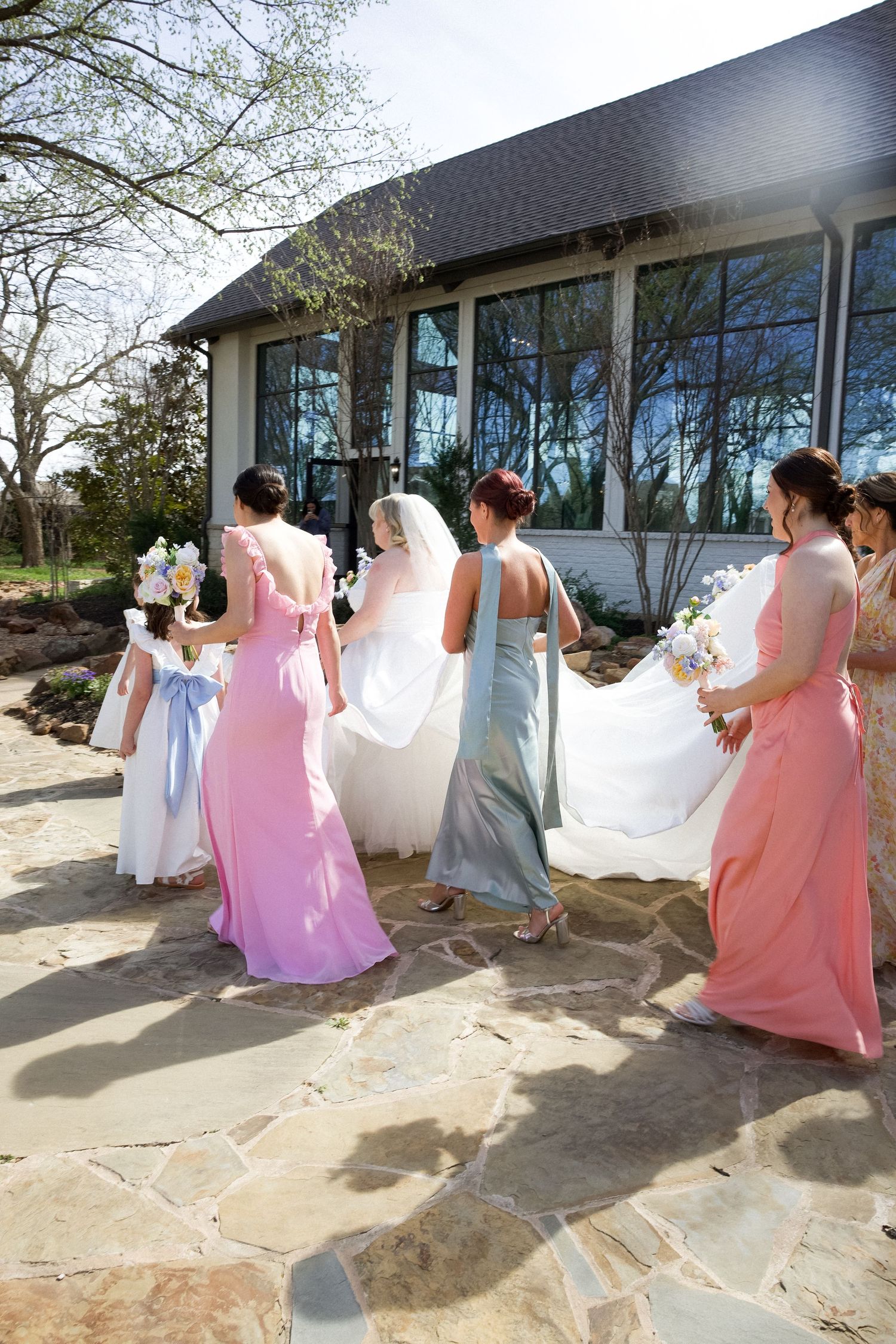 Bride in white veil walks with bridesmaids in pink and blue gowns along stone path to wedding chapel.