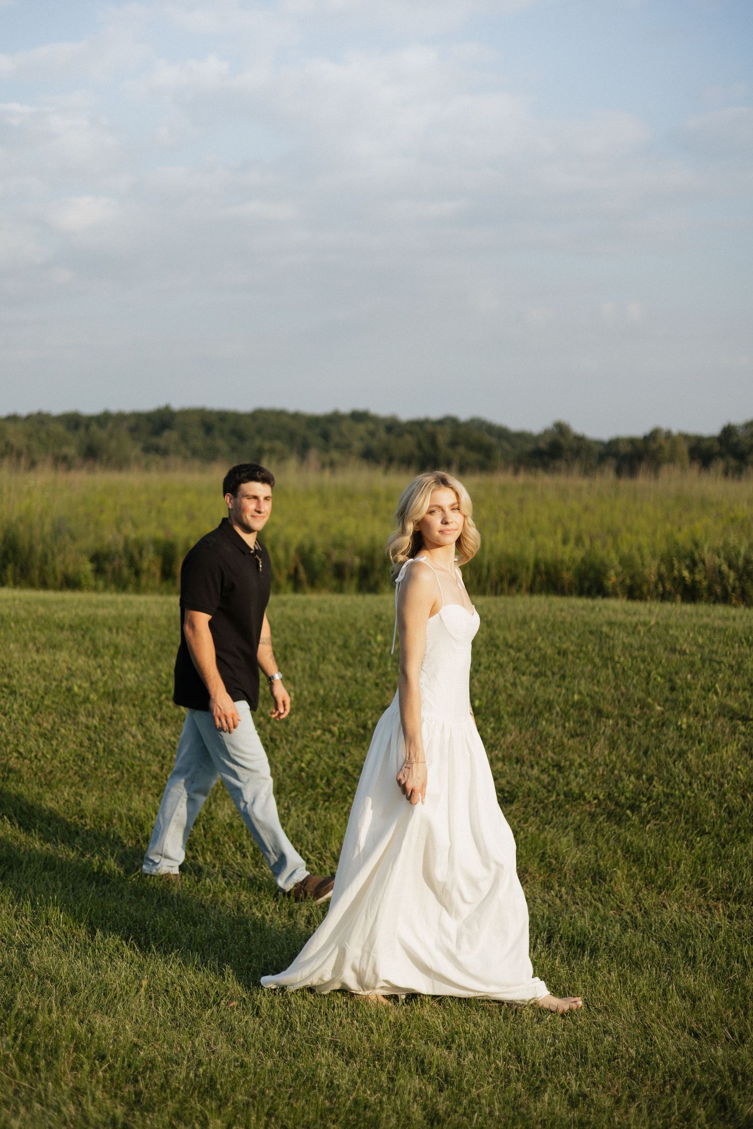 Bride in white gown walks with groom in black shirt across green field during golden hour outdoor wedding.