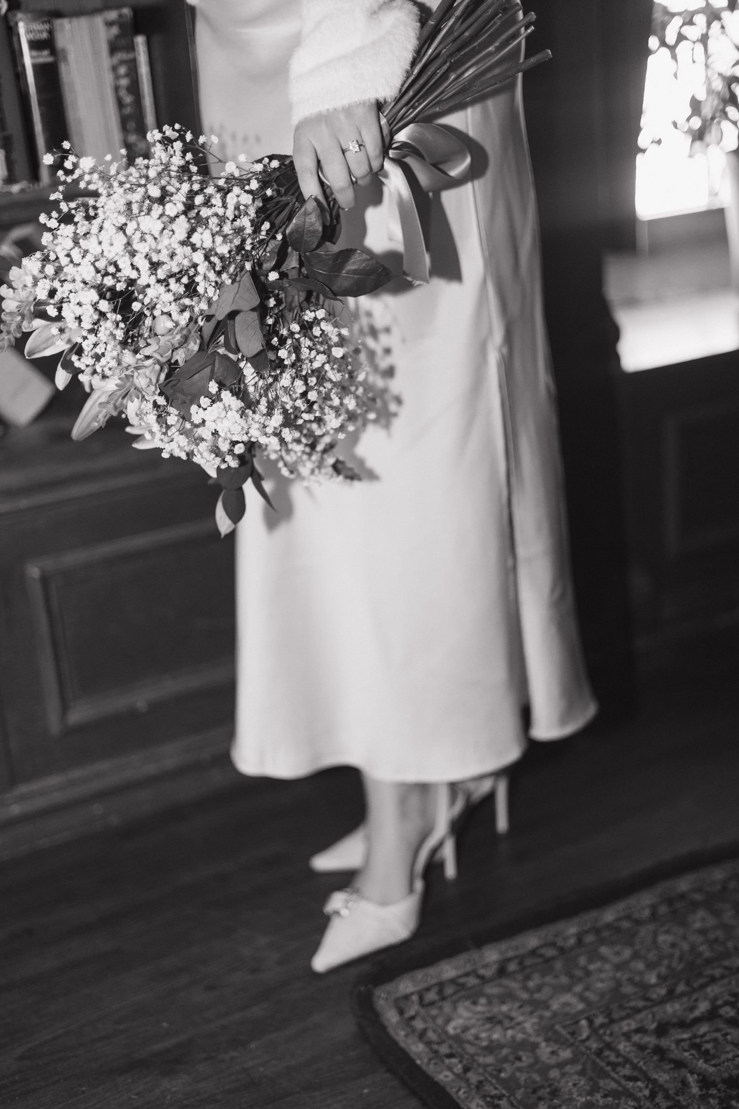 Black and white photo of a bride holding a baby's breath bouquet with ribbon, wearing a midi dress and bow heels.