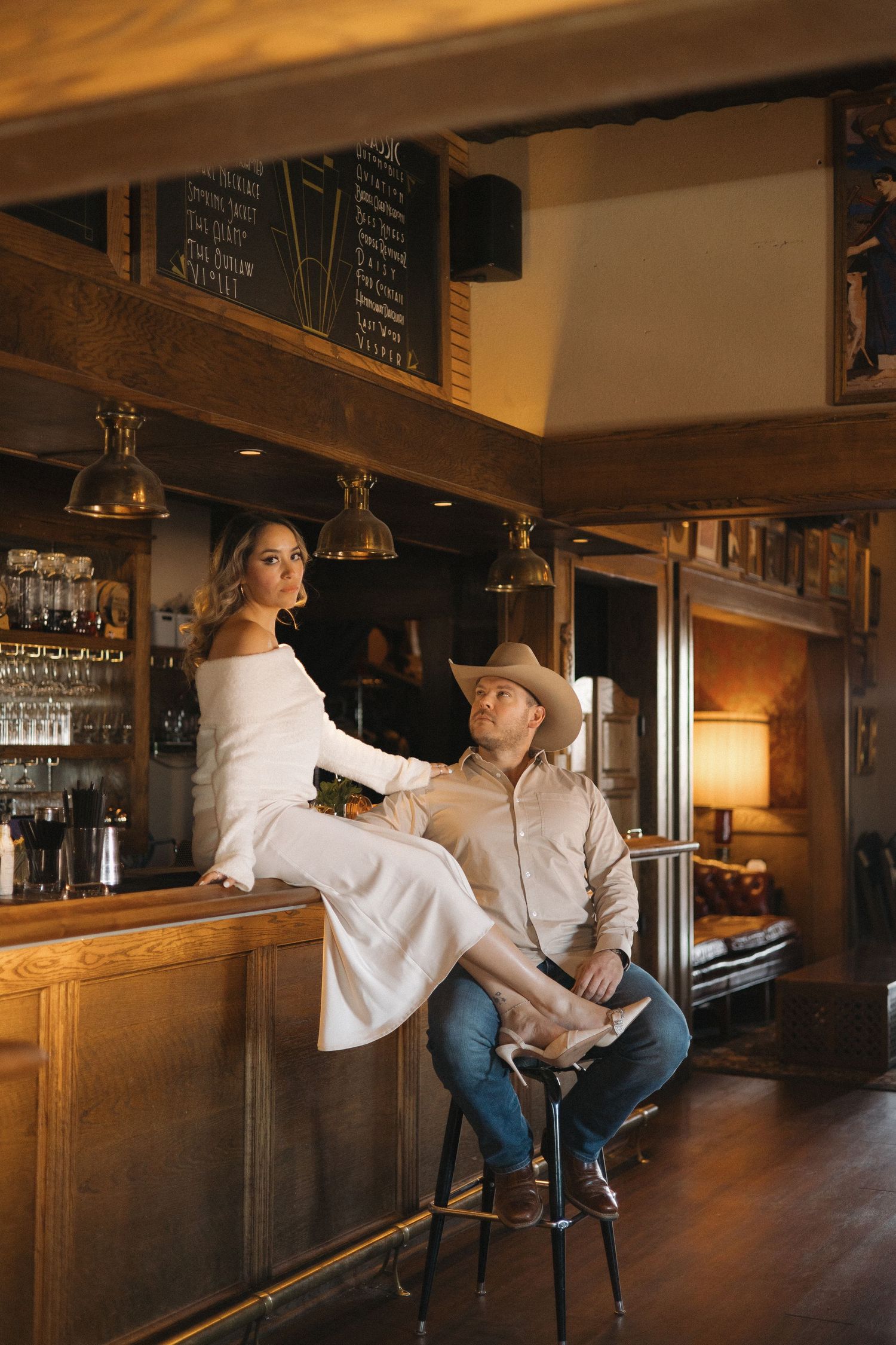 A couple poses in a rustic Western bar, woman in white dress on counter, man in cowboy hat seated on barstool.