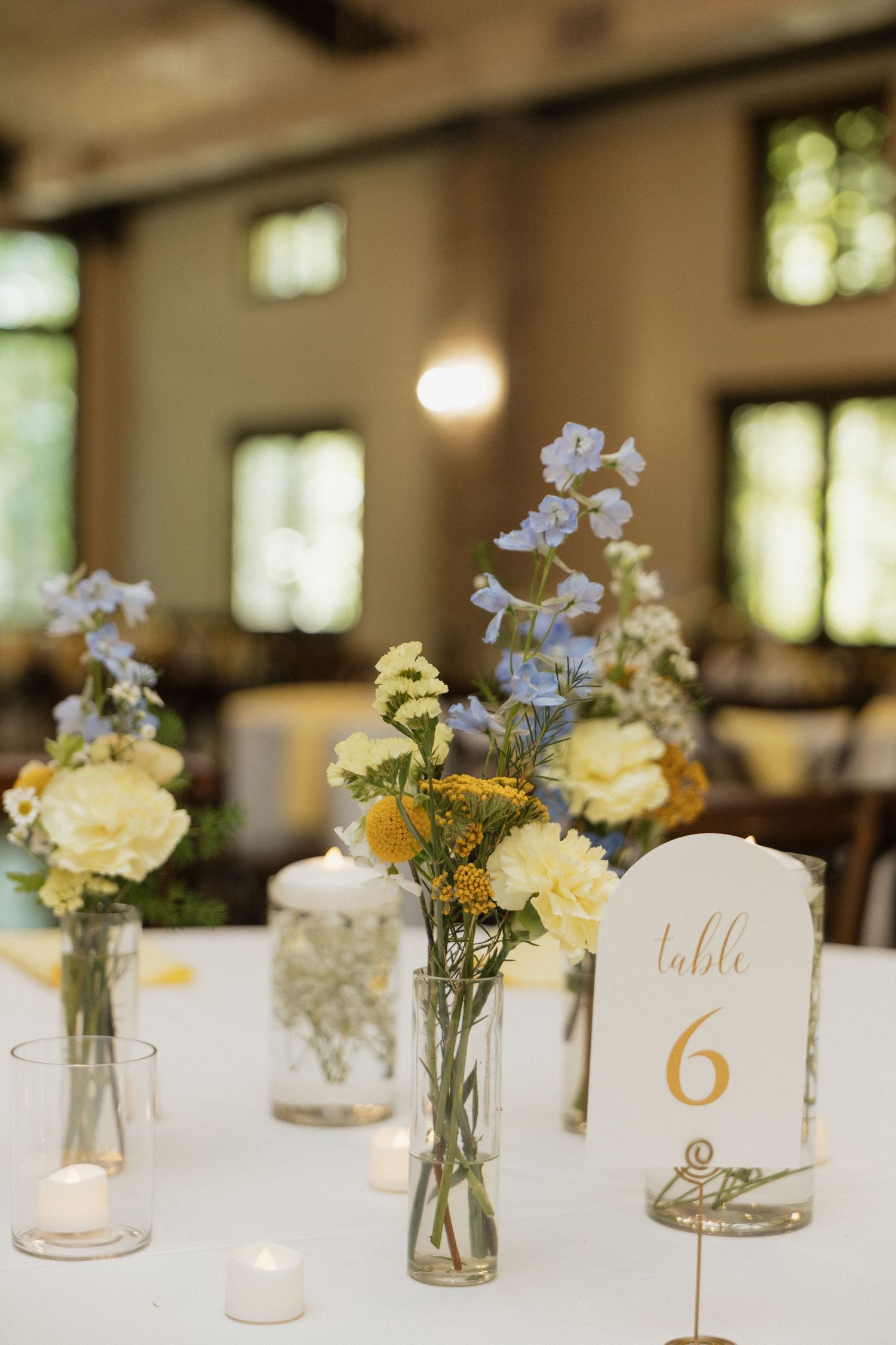 Wedding table 6 centerpiece with yellow, blue wildflowers in glass vases, candles, and elegant table number card.