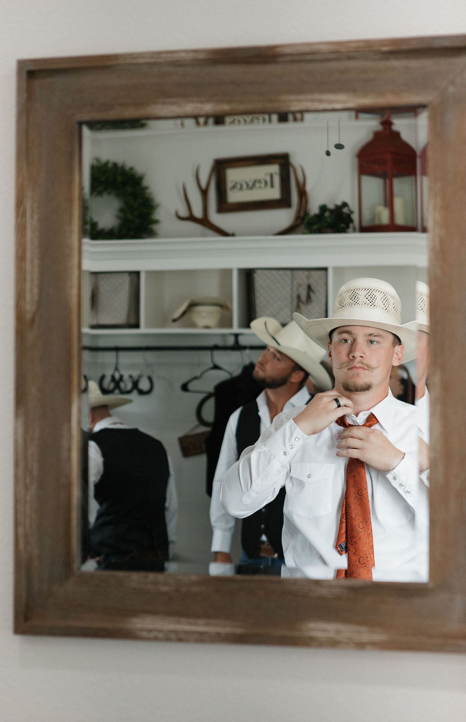 Person adjusting tie while looking in rustic framed mirror with cowboy hat and western decor.