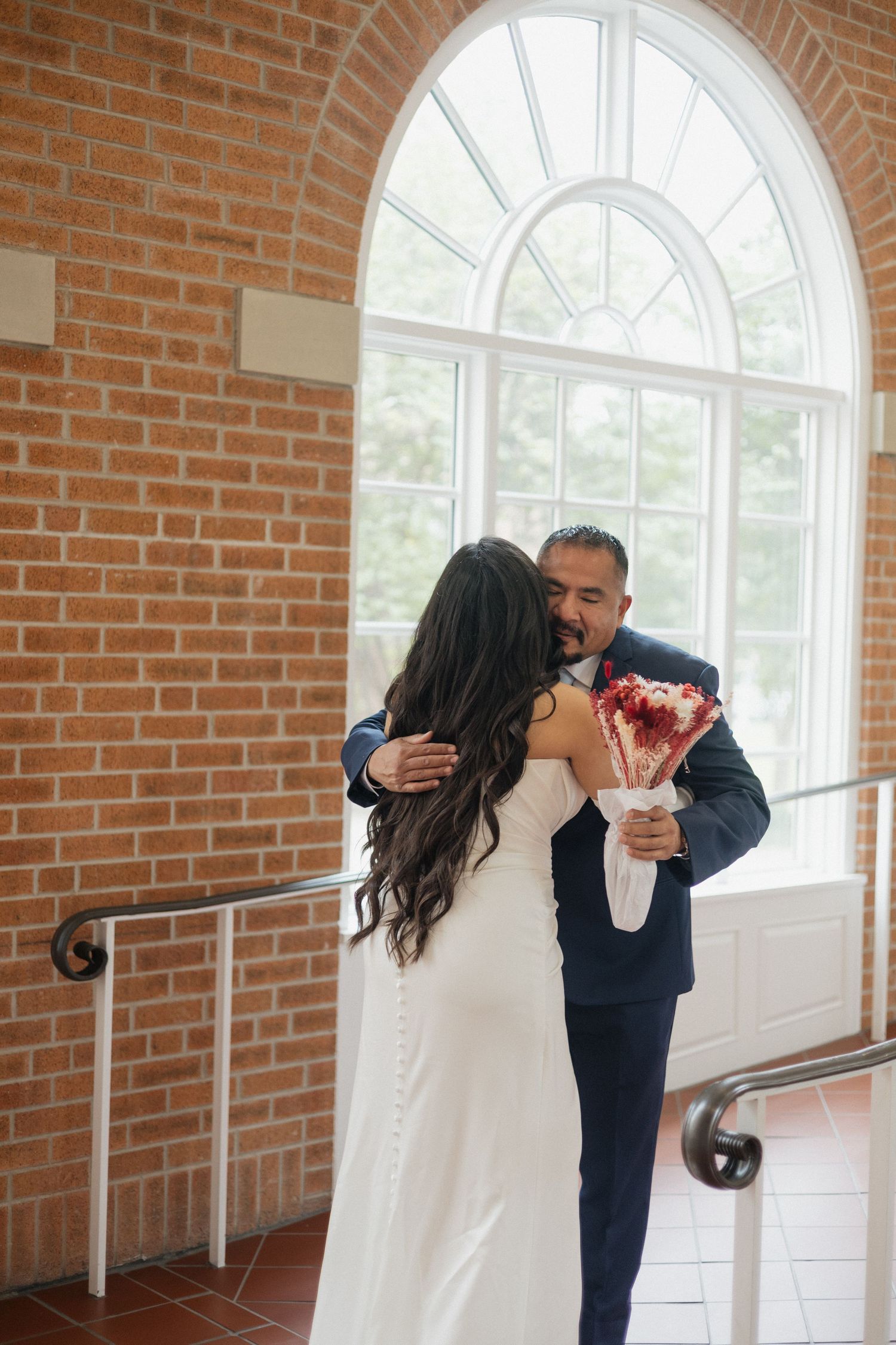 Bride and groom share a romantic embrace near an arched window in a brick venue, as groom holds red bridal bouquet.