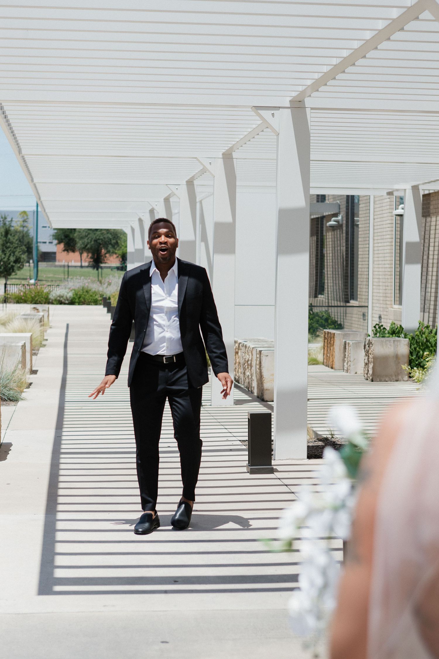 A person in a black suit walks under a modern white pergola structure on a sunny day.