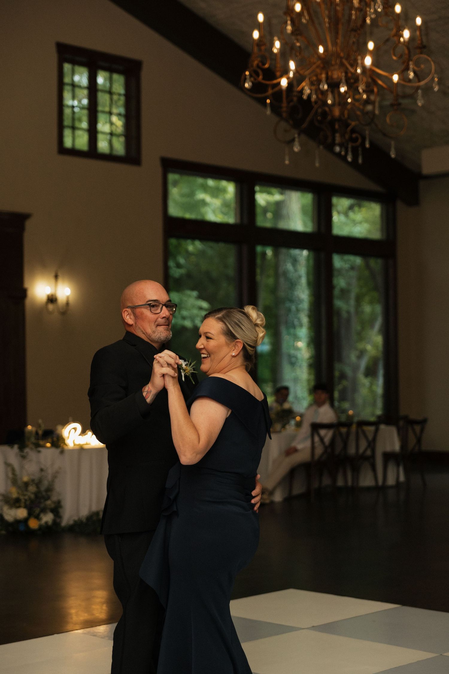 Couple in formal black attire share elegant dance beneath chandelier in warmly lit wedding reception.
