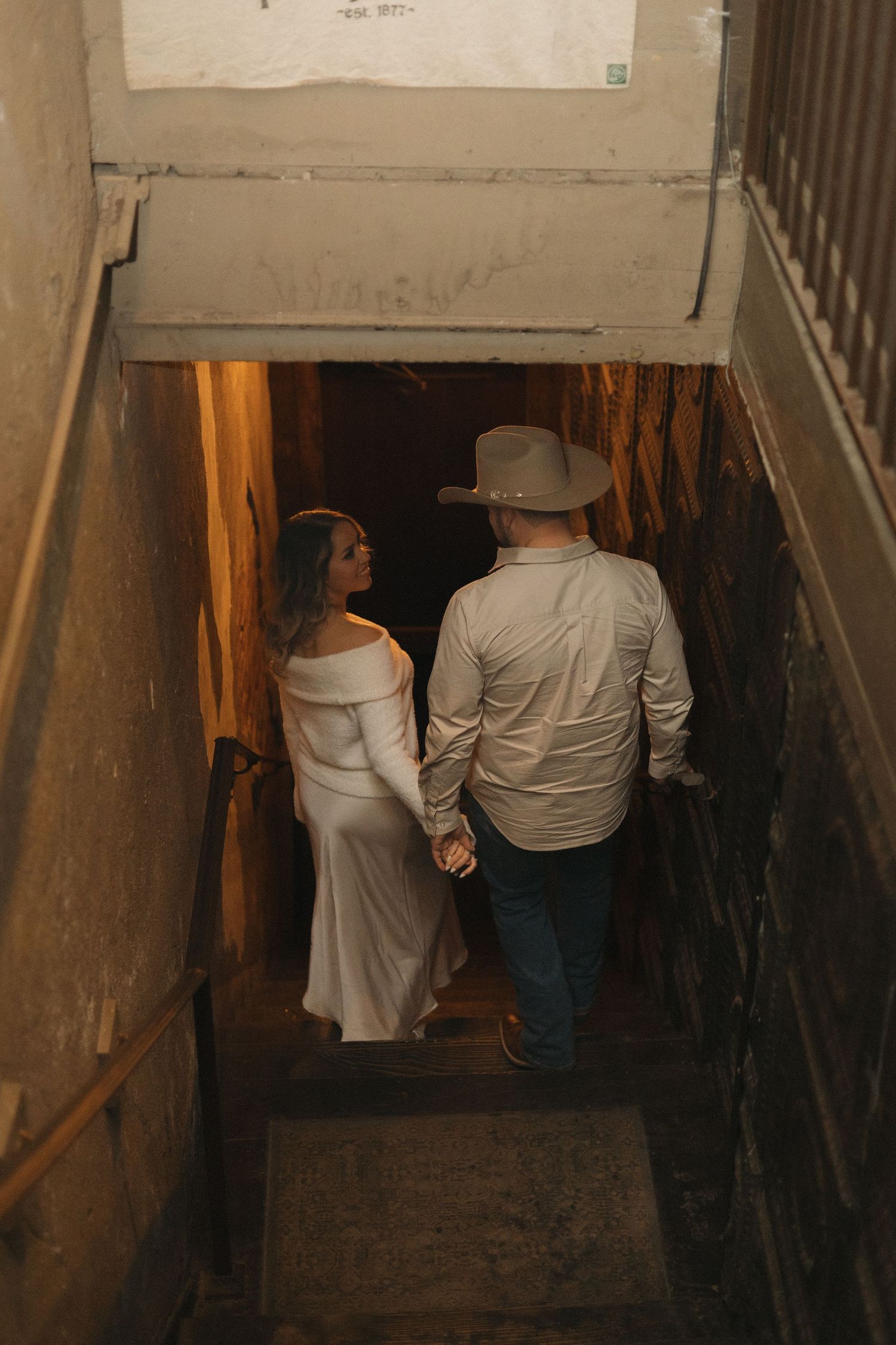 Couple holding hands descending rustic staircase, woman in white dress and man in cowboy hat.