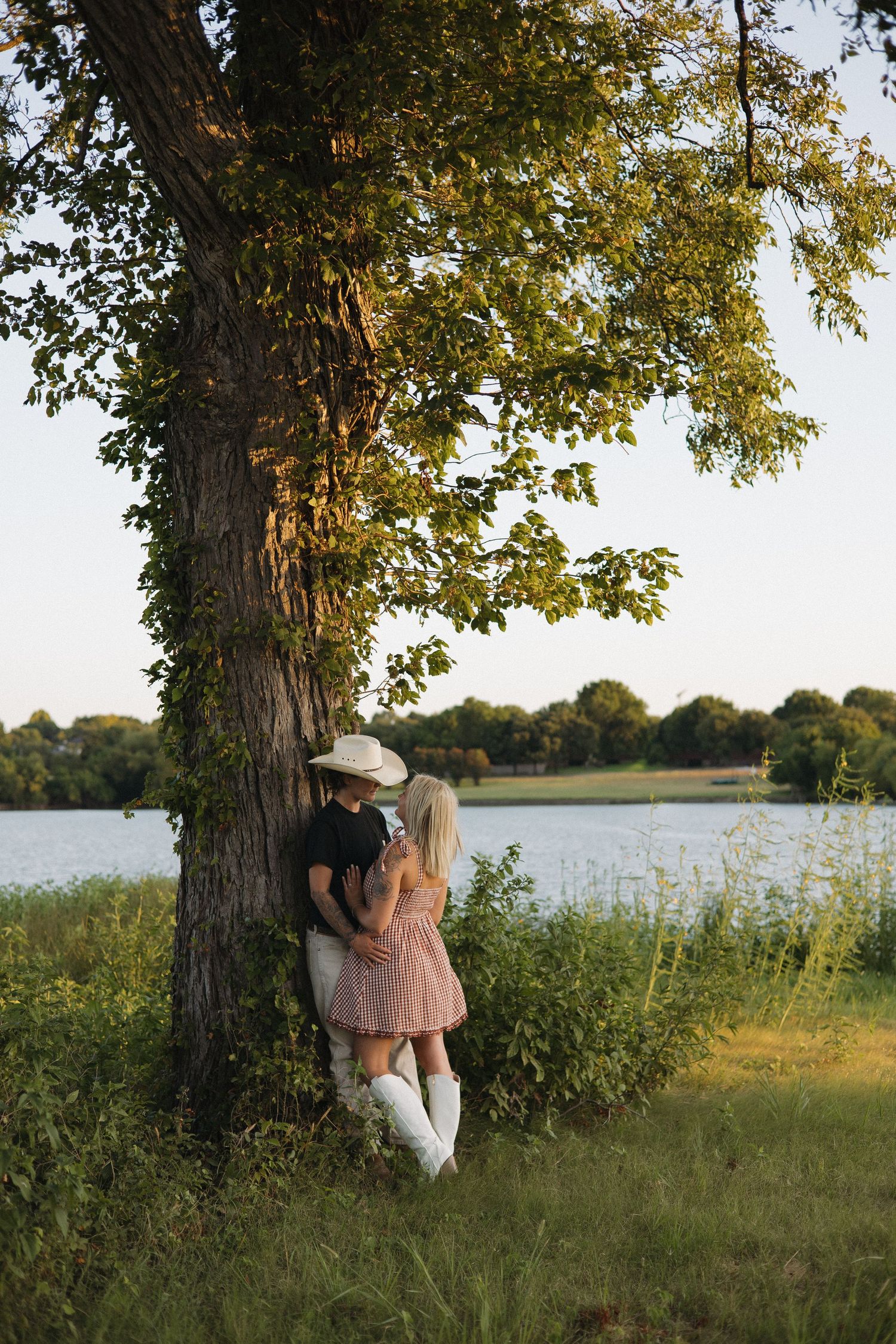 Couple in cowboy hats sharing a romantic moment by a large tree near a lake on a sunny day.