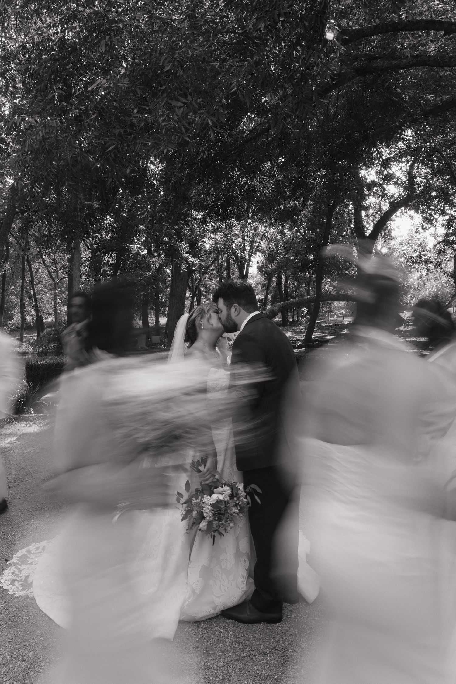Black and white photo of bride and groom kissing surrounded by blurred wedding guests in an outdoor garden setting.