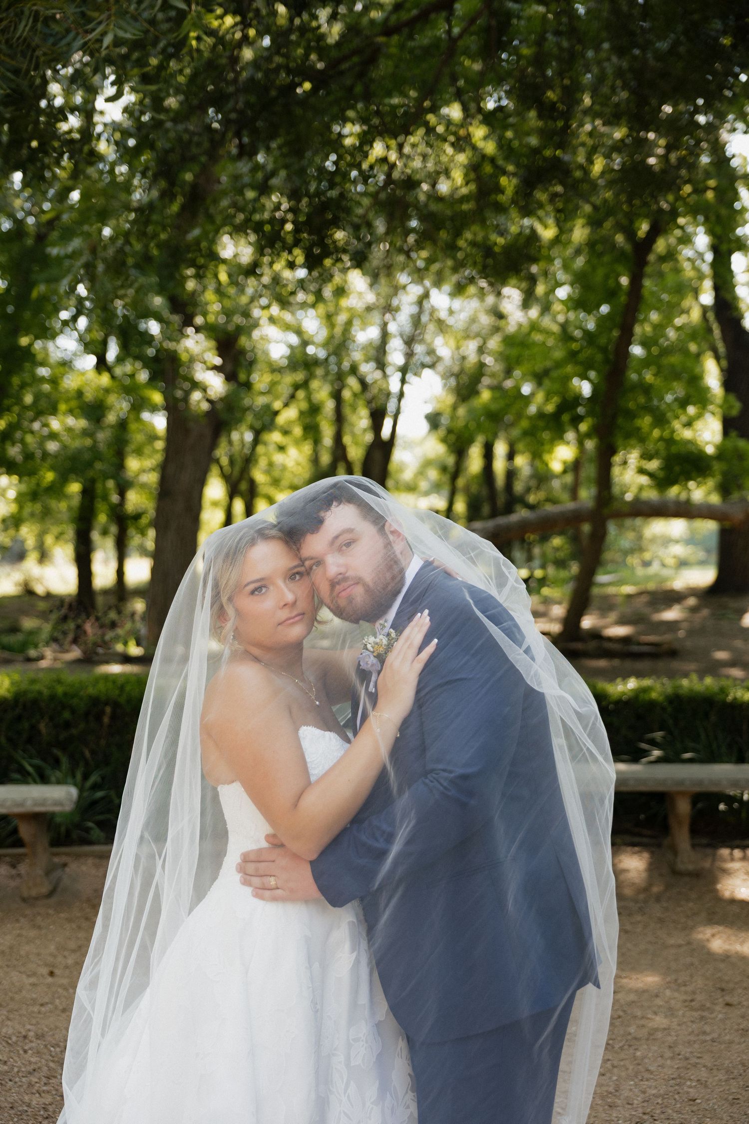 Bride and groom share a romantic moment under a flowing white veil in a lush green park setting.