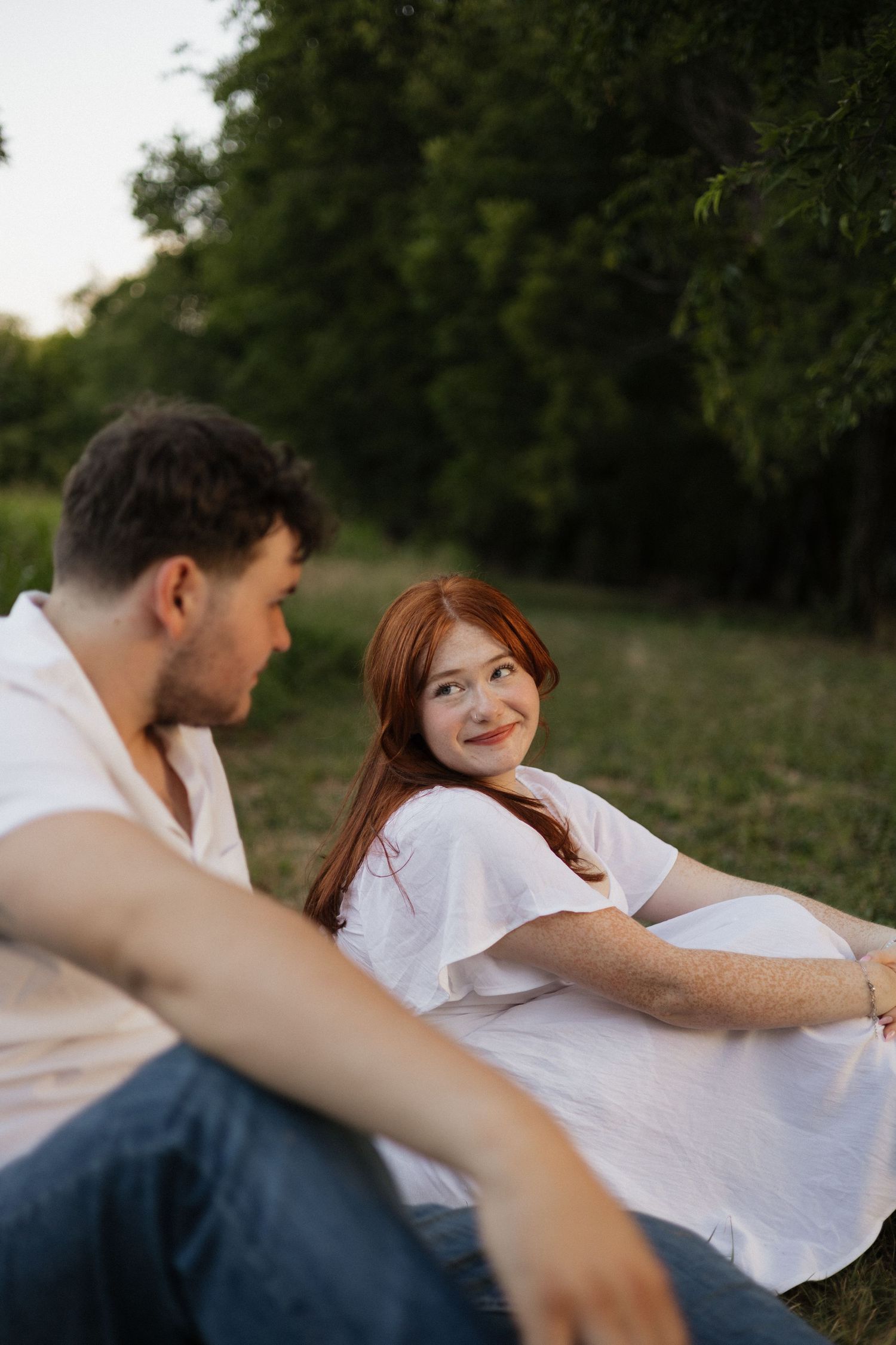 A pregnant woman in a white dress smiles at a man in a park setting during golden hour.