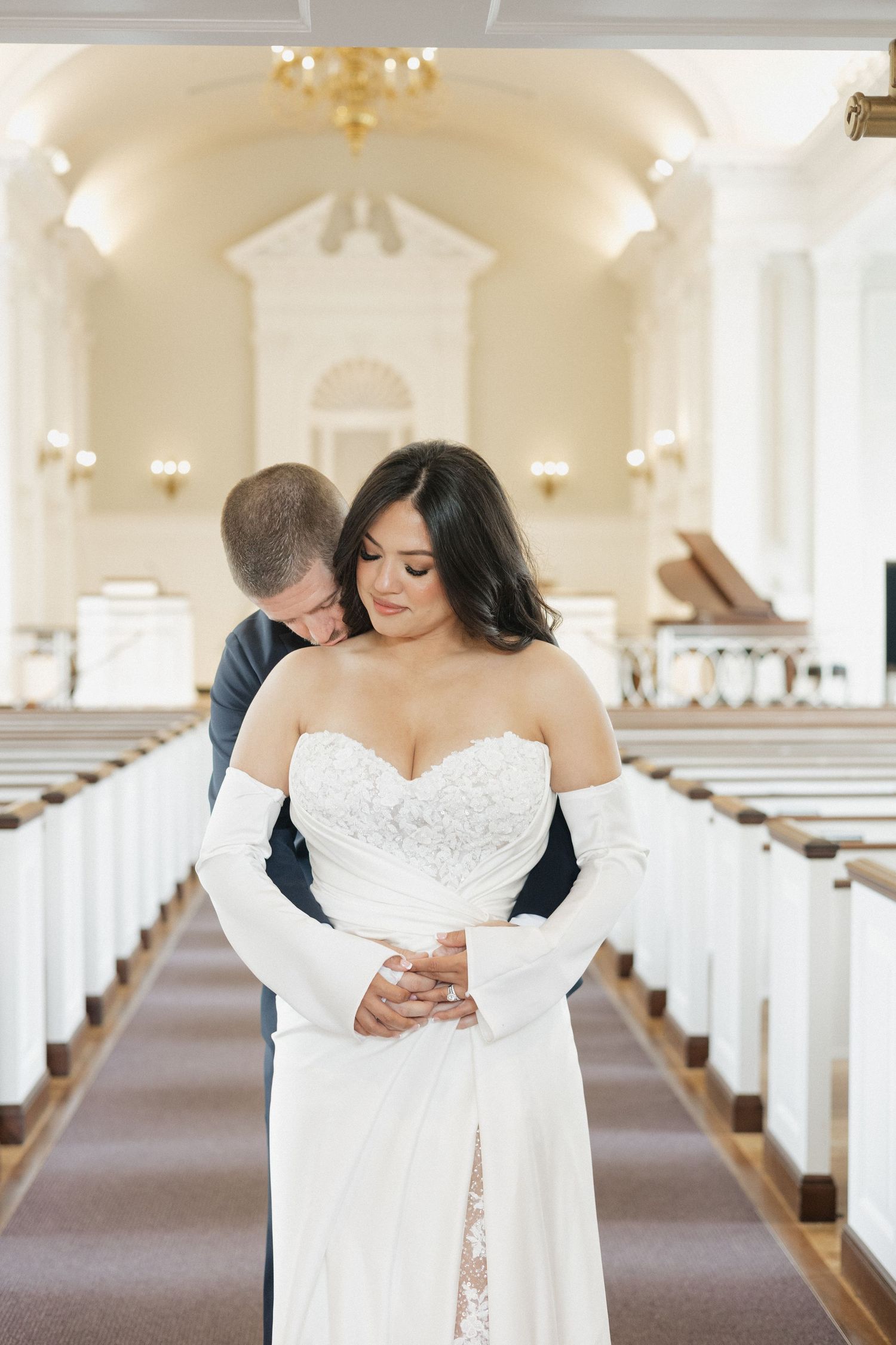 Groom embracing bride from behind in a classic white church with wooden pews.