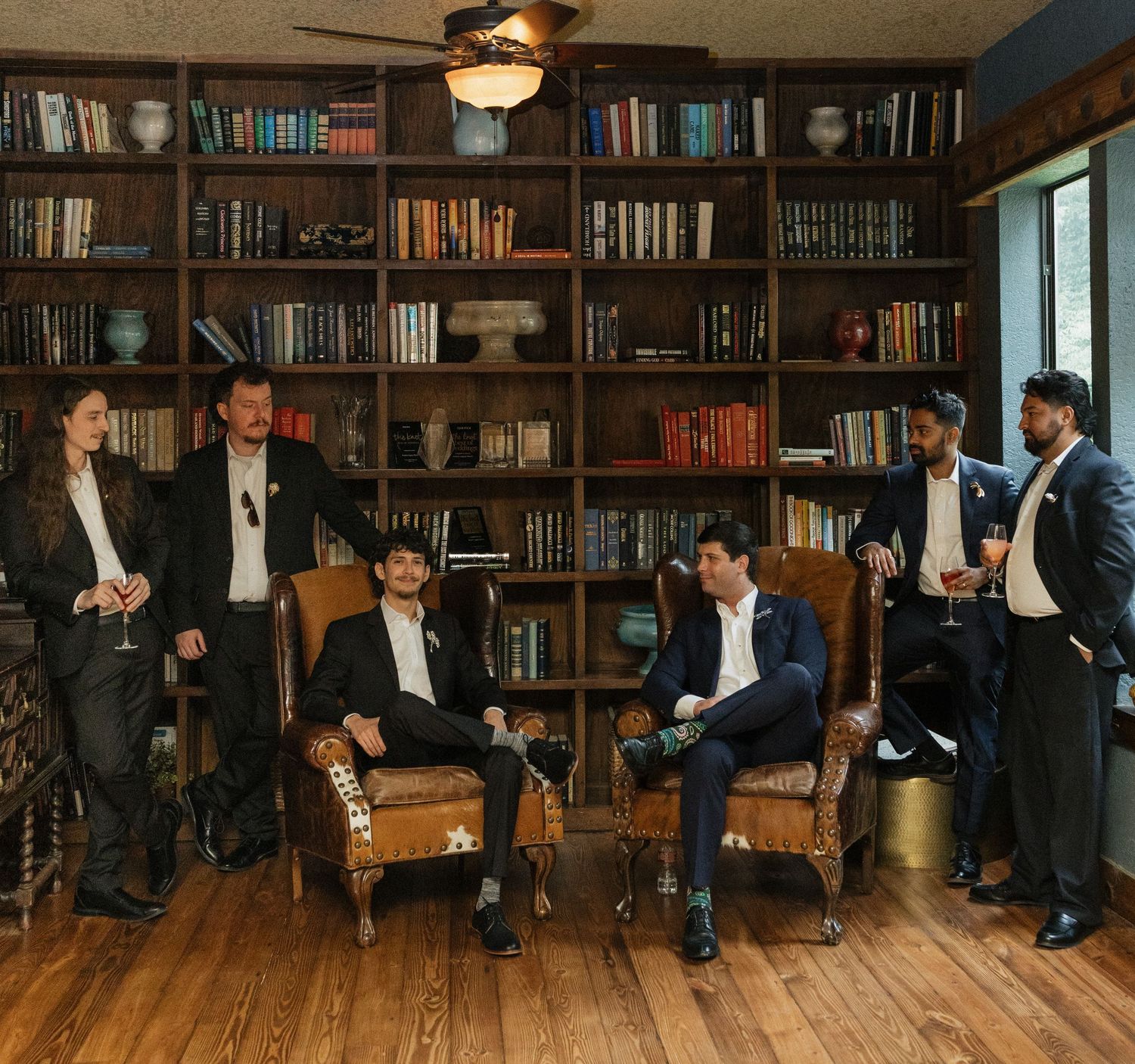 Six well-dressed men in suits pose in an elegant library with floor-to-ceiling bookshelves at a formal event.