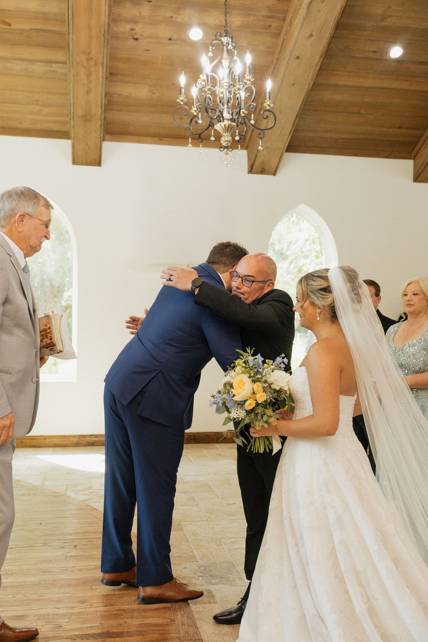 Groom in navy suit embraces father of bride during emotional wedding ceremony in white chapel.