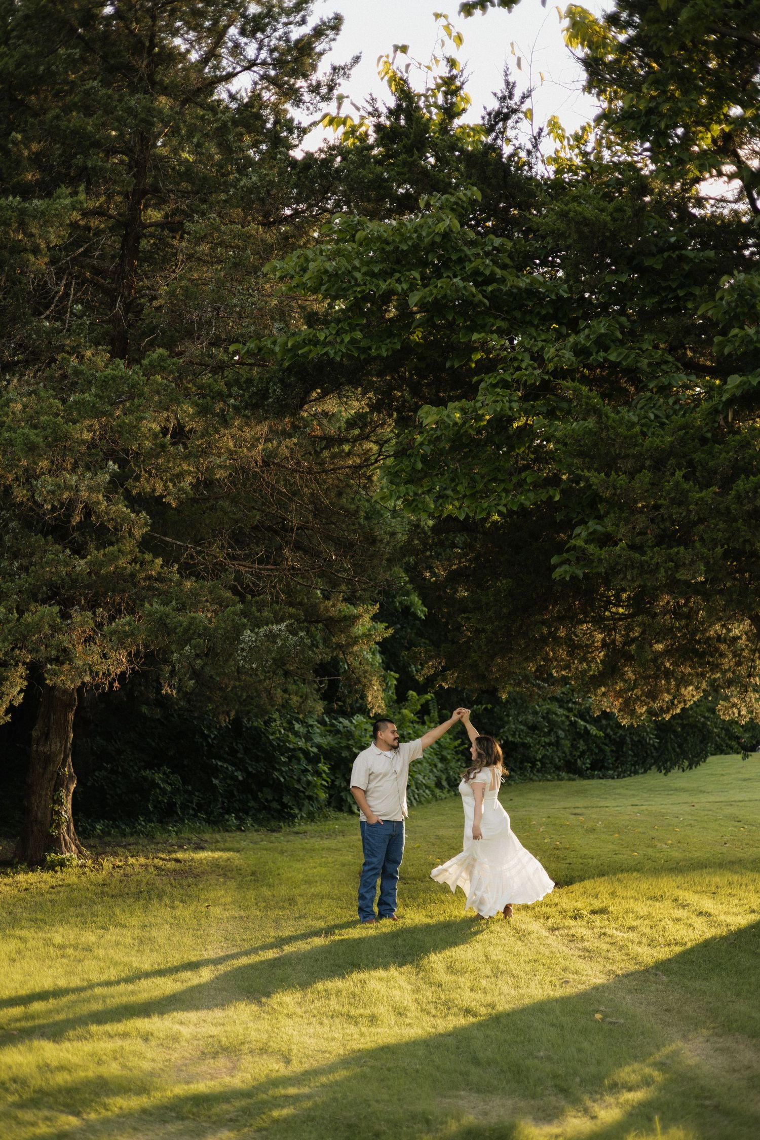 Couple dancing together on lush green grass surrounded by tall trees during golden hour.