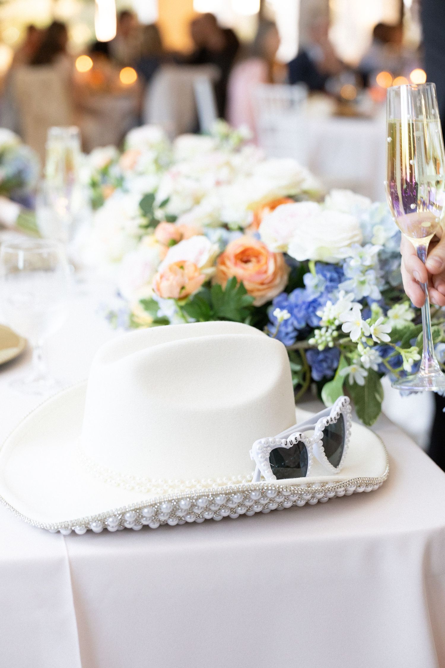 White cowboy hat with heart sunglasses on wedding reception table with floral centerpiece and champagne glass.