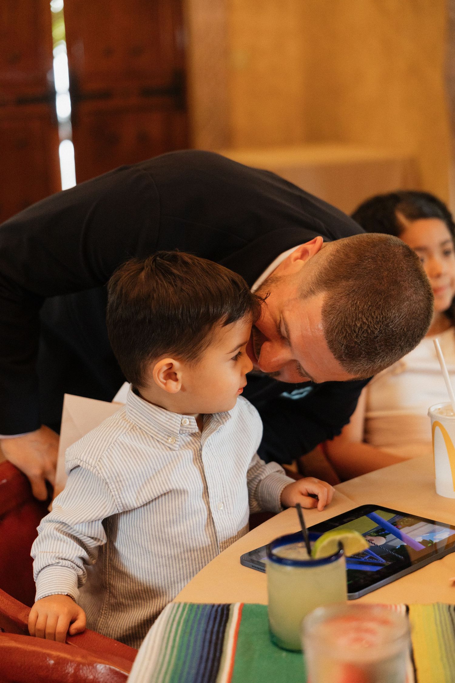 Father tenderly kissing young boy on forehead at restaurant table with tablet and colorful drinks nearby.
