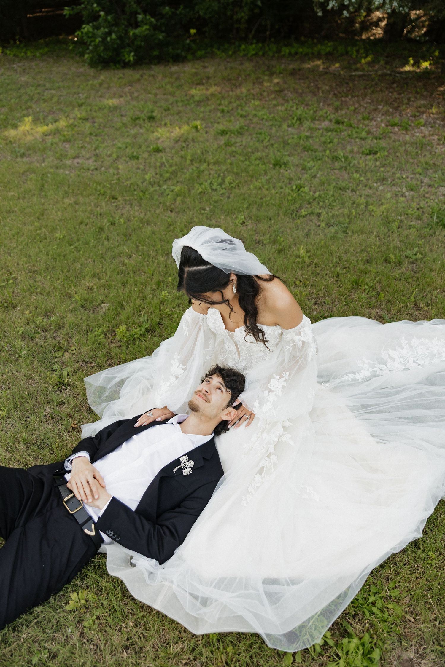 Bride in white lace gown kisses groom lying on grass during outdoor wedding photo session.